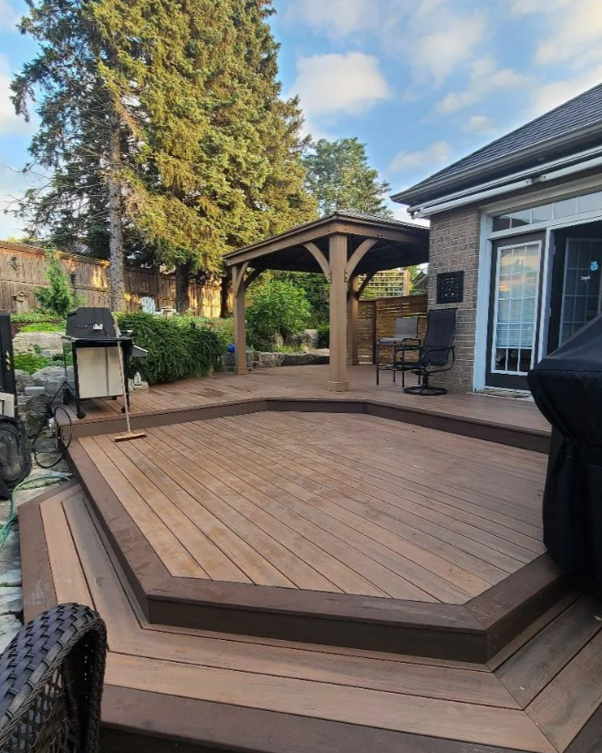 A backyard deck with a divided level, wooden furniture, a small gazebo, and surrounding greenery.