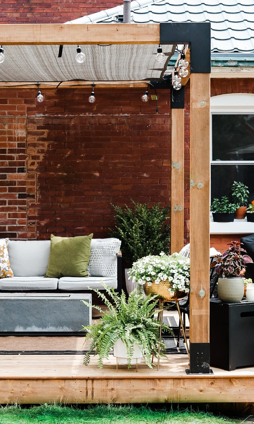 Outdoor patio area with string lights, a brick wall, a white couch with pillows, and various potted plants on a raised wooden deck.