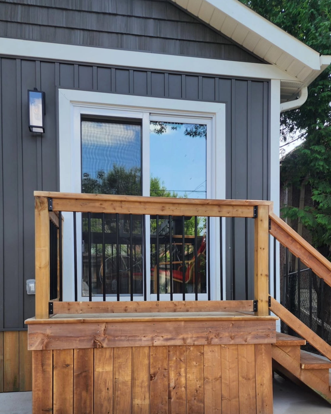 View of a small wooden deck with a railing, attached to a house with dark gray siding and a sliding glass door, leading into a room with a red chair inside. There is a window on the house, a light fixture on the wall, and trees visible in the reflection and background.