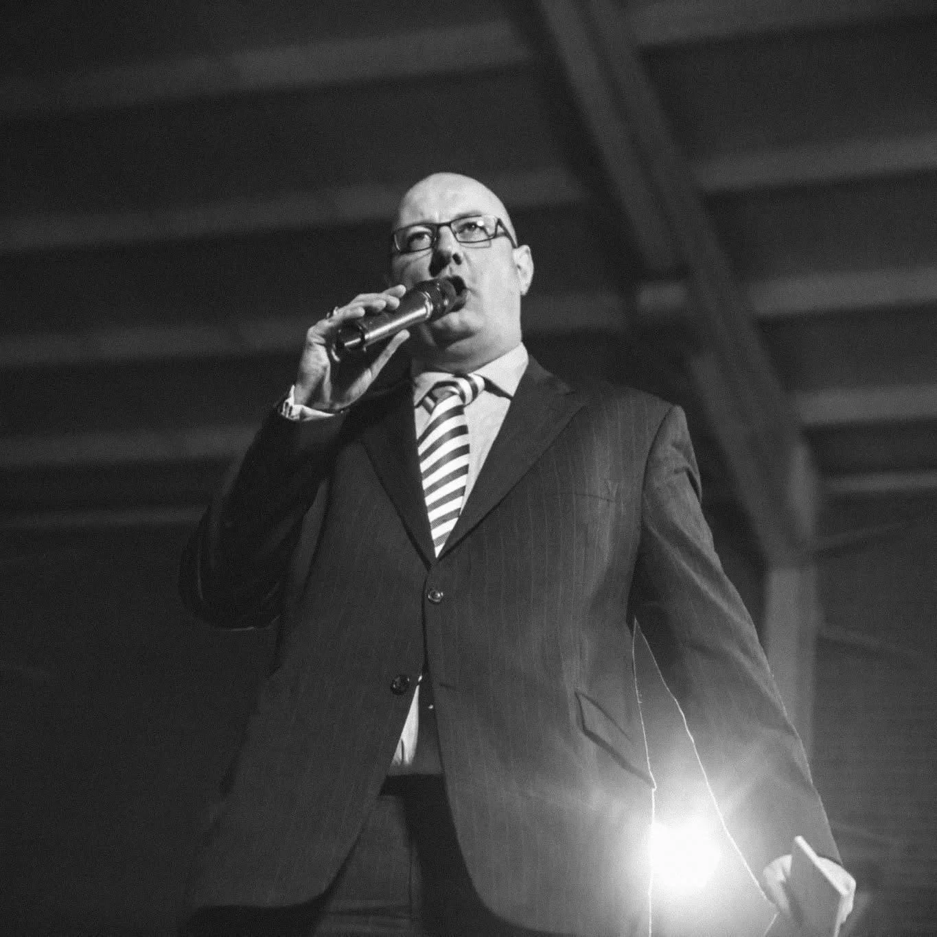 A black and white photo of a man in a suit and striped tie, wearing glasses, holding a microphone, speaking in front of an overhead structure with dramatic lighting from below.