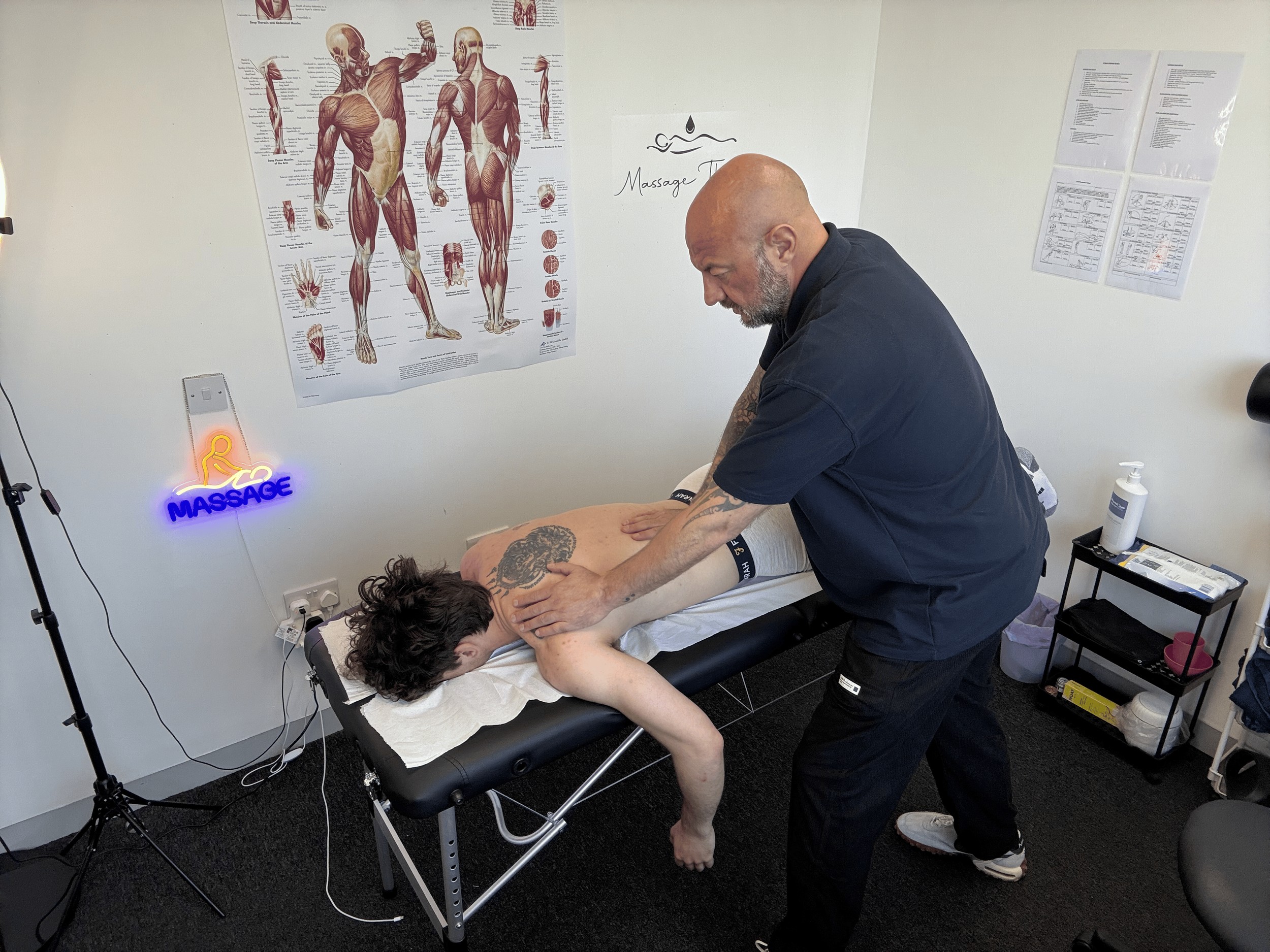 A massage therapist is giving a massage to a male client lying face down on a massage table, in a room with anatomical posters and a neon sign that says 'Massage' on the wall.