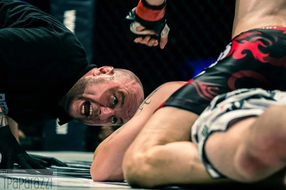 Mixed martial arts fight with one fighter on the ground and the other fighter leaning over. The fighter on the ground appears to be in distress. The fighter leaning over has a clenched fist, indicating a victory or dominance.