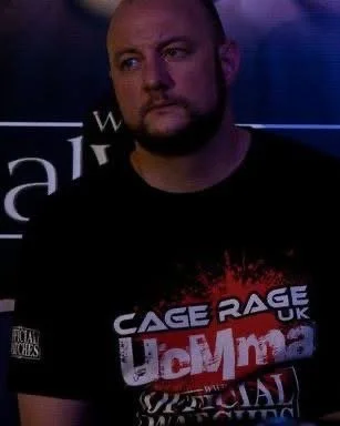 A man with a beard looks to the side in a dimly lit setting, wearing a black t-shirt with red and white text related to MMA or cage fighting.