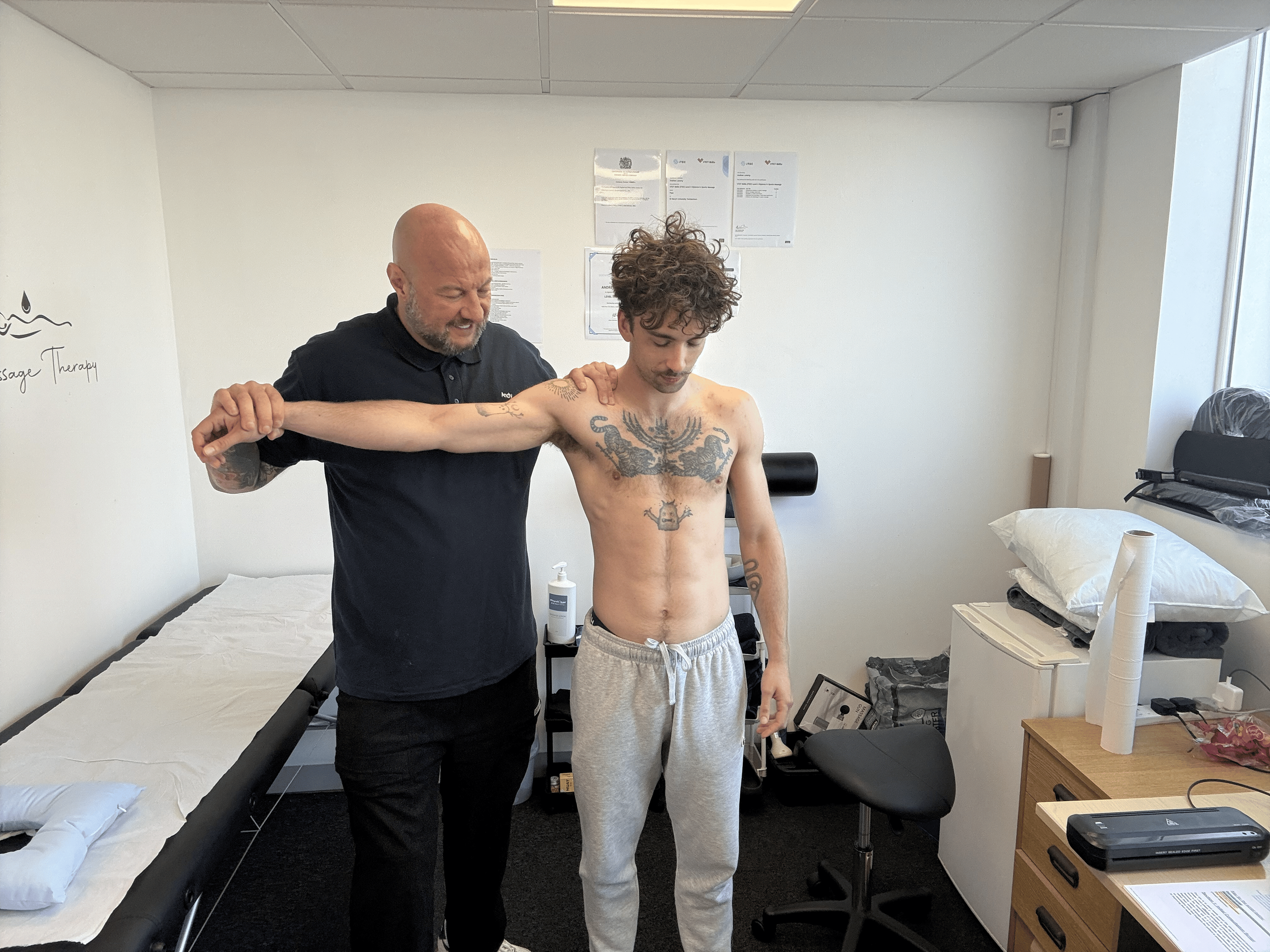 A man with curly hair and tattoos on his chest and arm is standing in a medical examination room, with a medical professional assessing his shoulder. The room has a bed with white sheets, a few chairs, medical supplies, and some medical posters on th