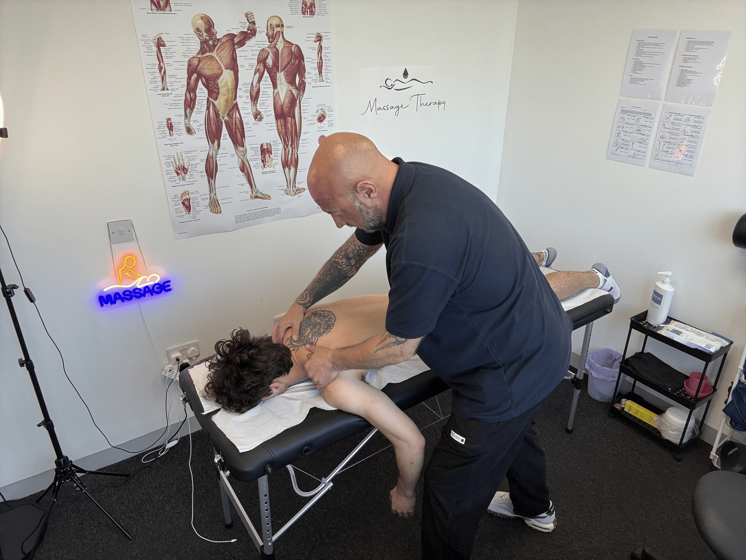Massage therapist giving a massage to a person lying face down on a massage table in a room with anatomical charts on the wall.