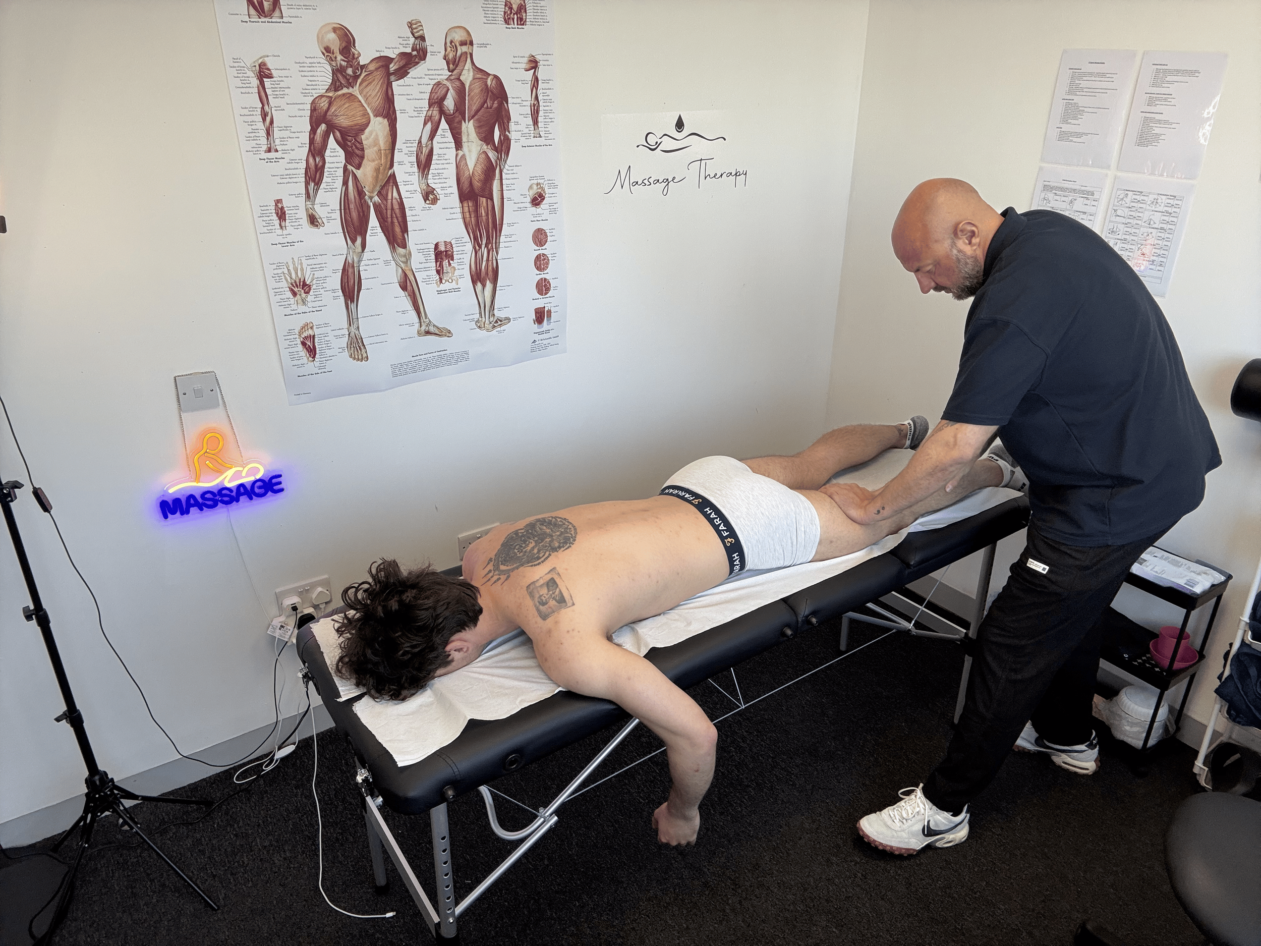 Massage therapy session in progress in a wellness center. A massage therapist is working on a client lying face down on a massage table, with a tattoo visible on their back. The room has anatomical posters on the wall and a neon sign that says 'Massa