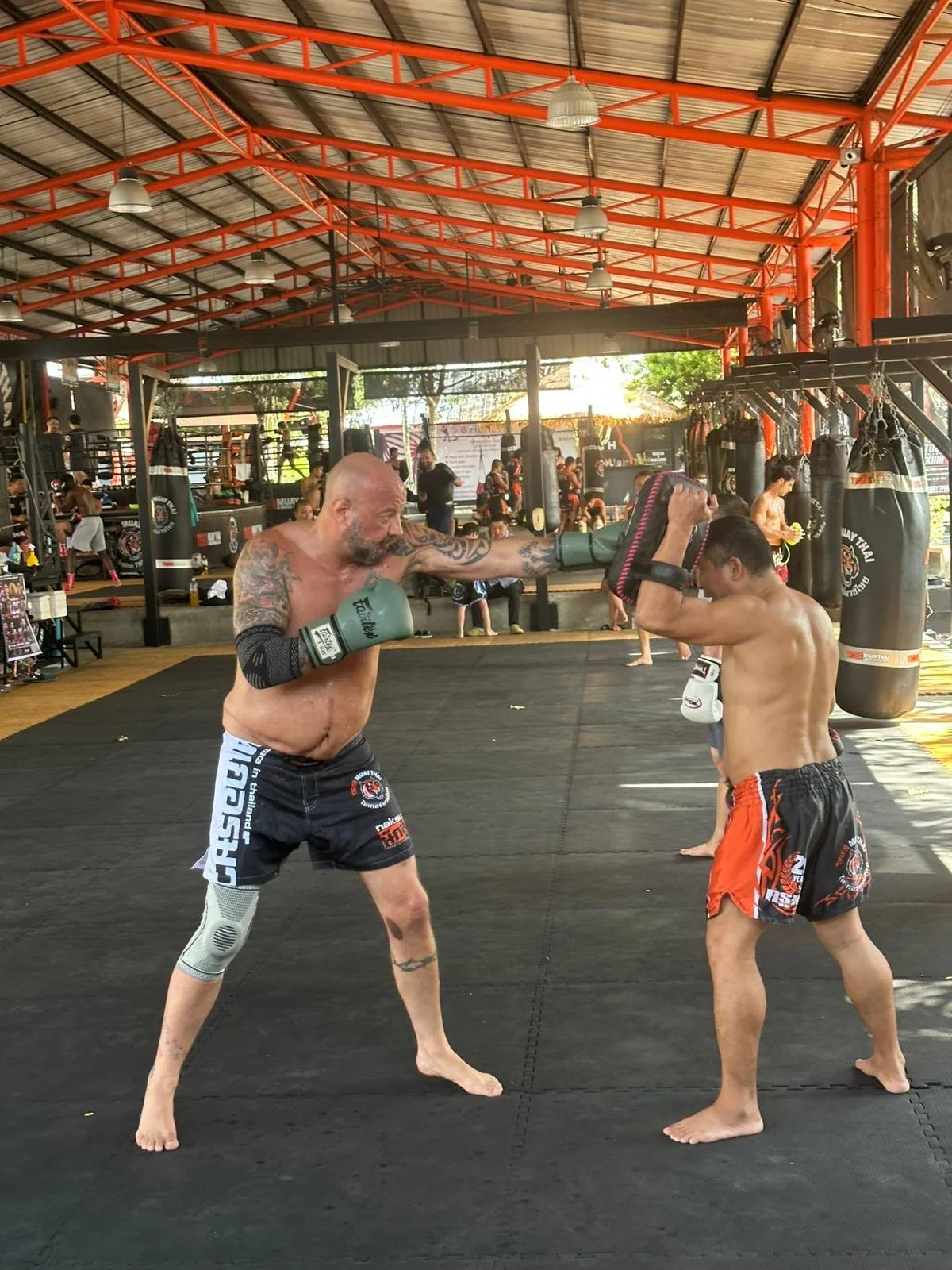 Two men practicing Muay Thai in a gym. The man on the left is throwing a punch with his right hand, while the man on the right is holding focus mitts. They are both shirtless and wearing shorts. The gym has punching bags hanging and other people trai