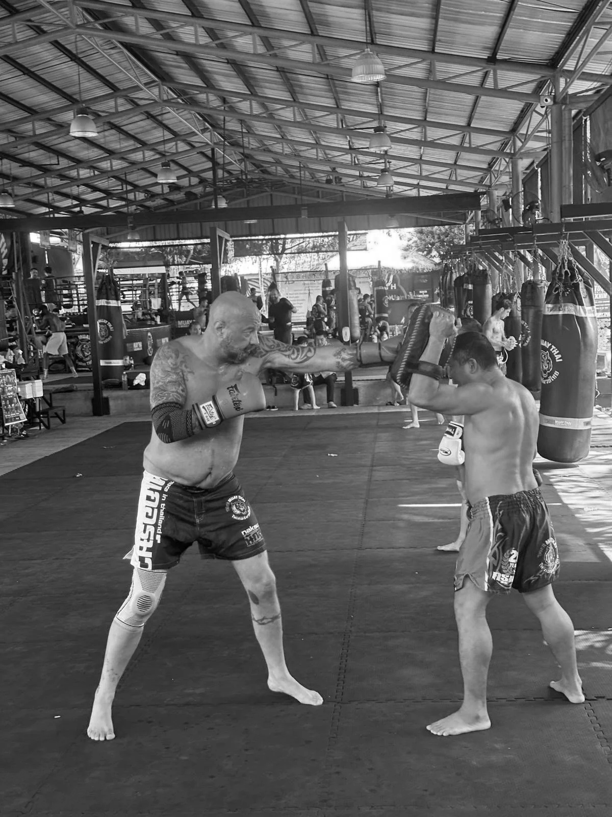 Two shirtless men with boxing gloves sparring in a gym. The man on the left has tattoos and is throwing a punch, while the man on the right is blocking with his gloves. The gym has punching bags and other people practicing in the background.