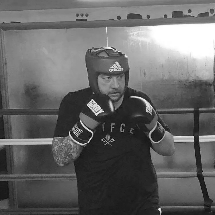 A man wearing boxing gloves and a headgear stands in a boxing gym, ready to train or spar.