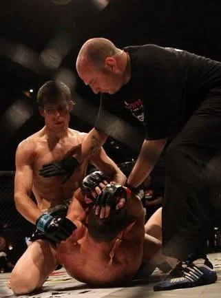 Mixed martial arts fight with referee checking on a fighter who is lying on the ground while the other fighter stands nearby.
