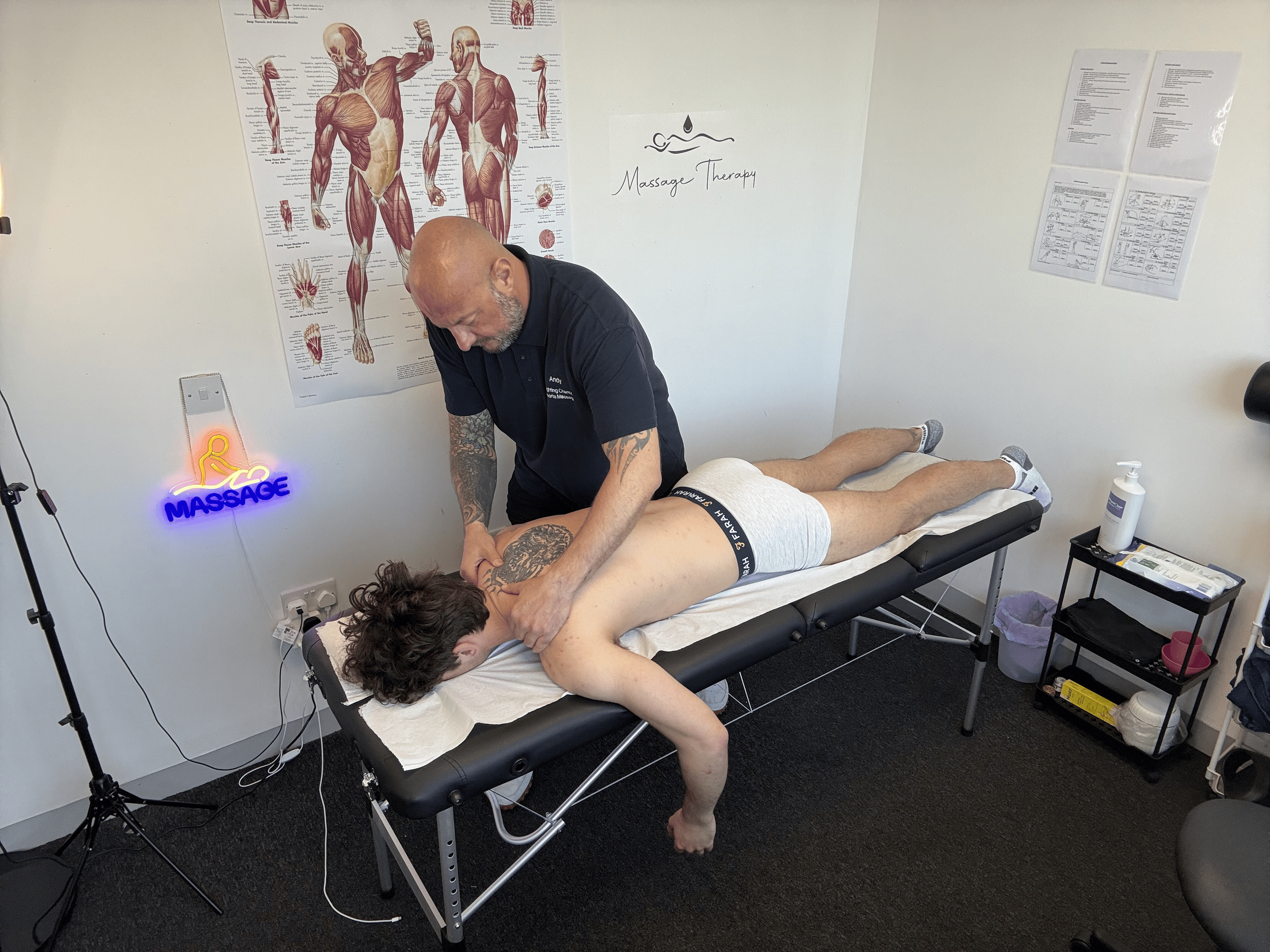 A massage therapist gives a massage to a man lying face down on a massage table in a clinic room with medical posters on the wall, including anatomical diagrams of muscles and nervous system.