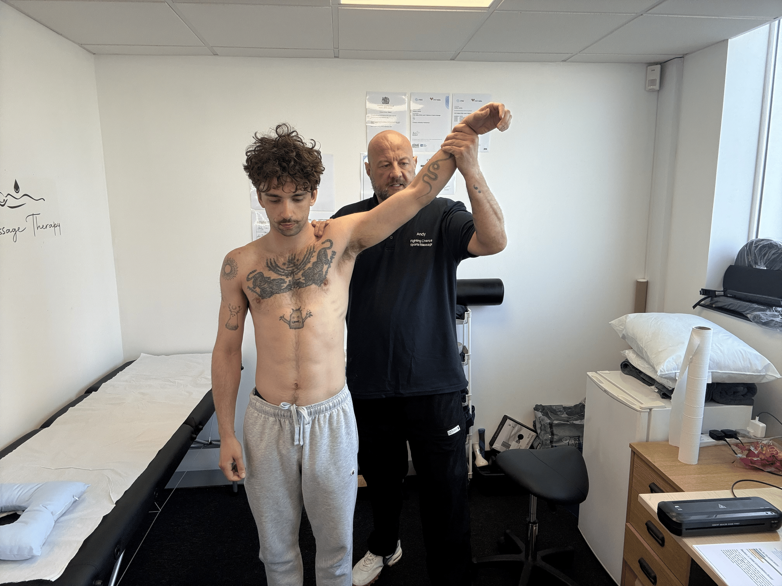A young man with curly hair and tattoos on his chest and arm is standing shirtless in a medical or massage therapy room, with a professional man holding his arm up and examining it.