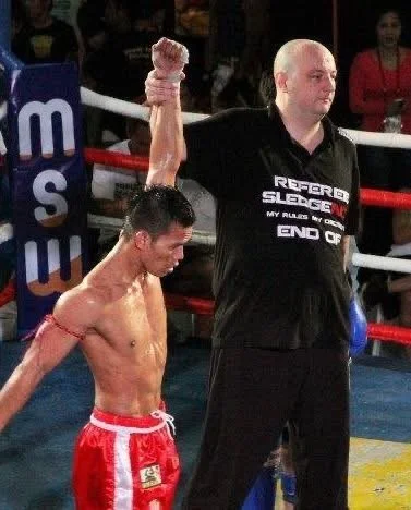 Muay Thai fighter with shaved head and red shorts with white trim standing with his arm raised by the referee in a boxing ring.