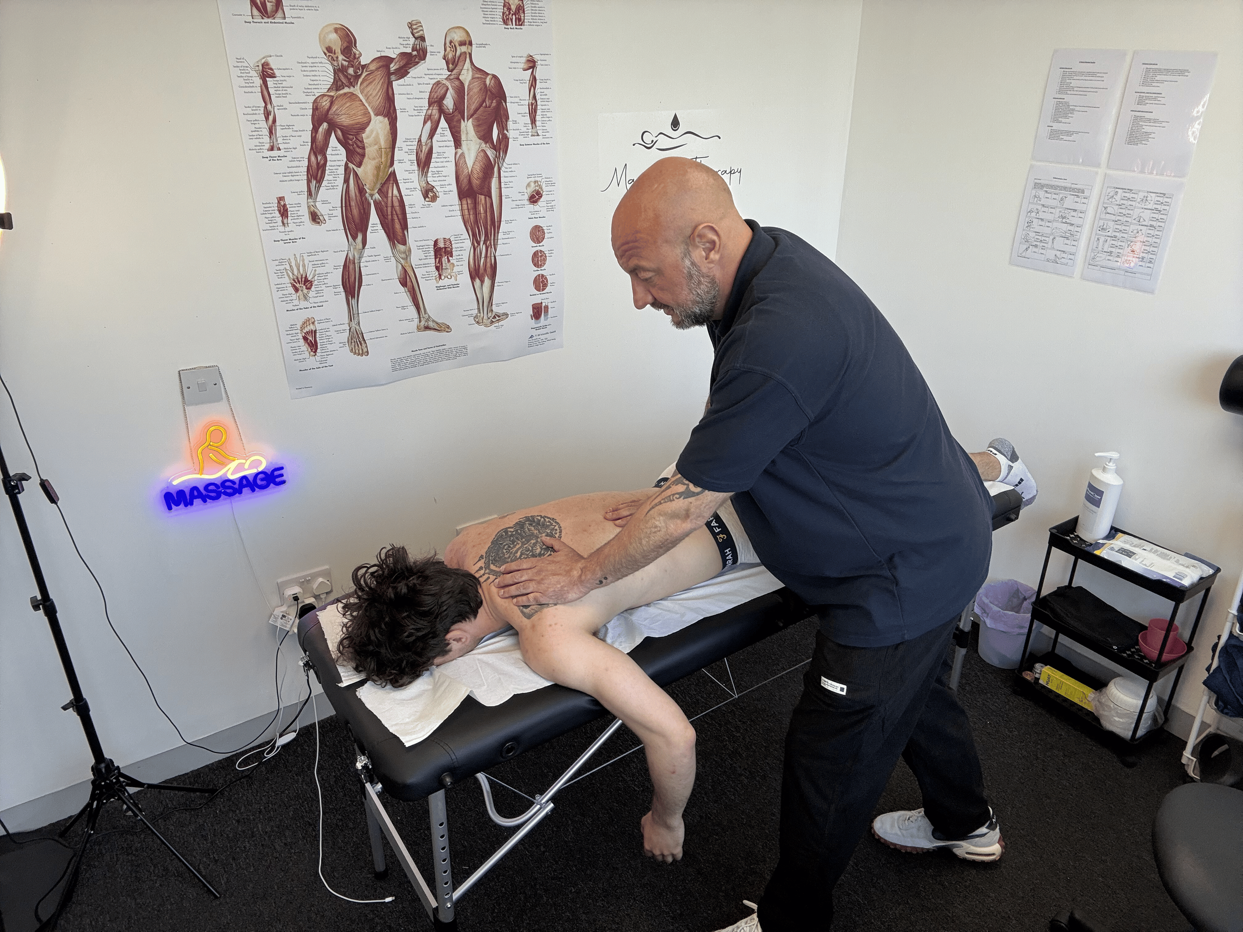 A massage therapist provides a massage to a person lying face down on a massage table in a therapy room. The room has anatomy posters on the wall and a small shelf with massage supplies.