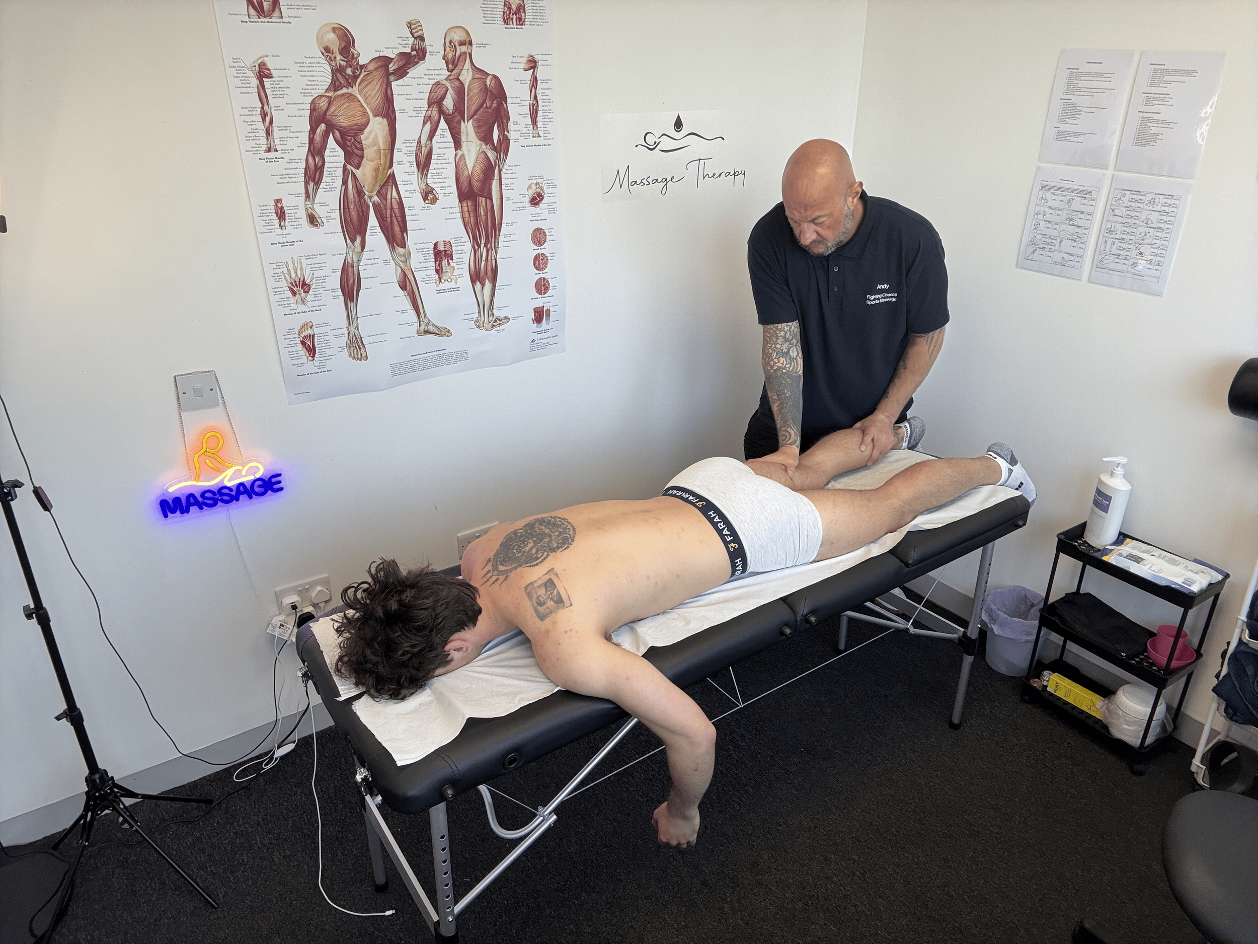A massage therapist is giving a massage to a man lying face down on a massage table. The room has anatomical posters on the wall, and a massage therapy sign is visible. The therapist is bald and wearing a black polo shirt. There is a neon sign that s