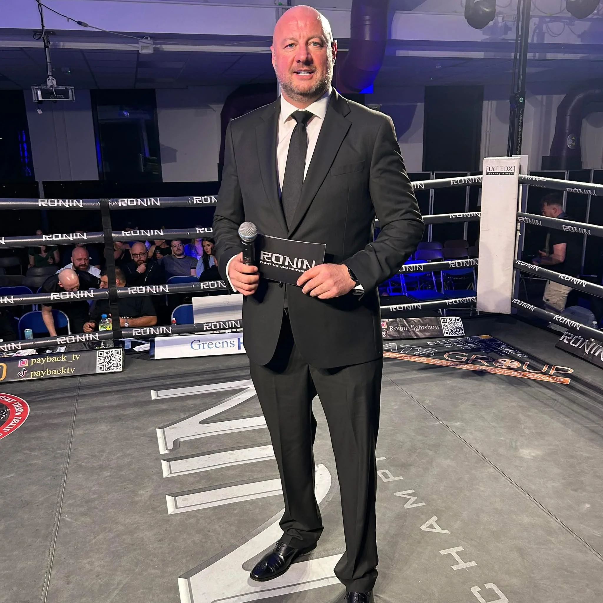 A man in a black suit and tie standing on stairs inside a boxing ring, holding a microphone and smiling. The background includes an audience watching the event.