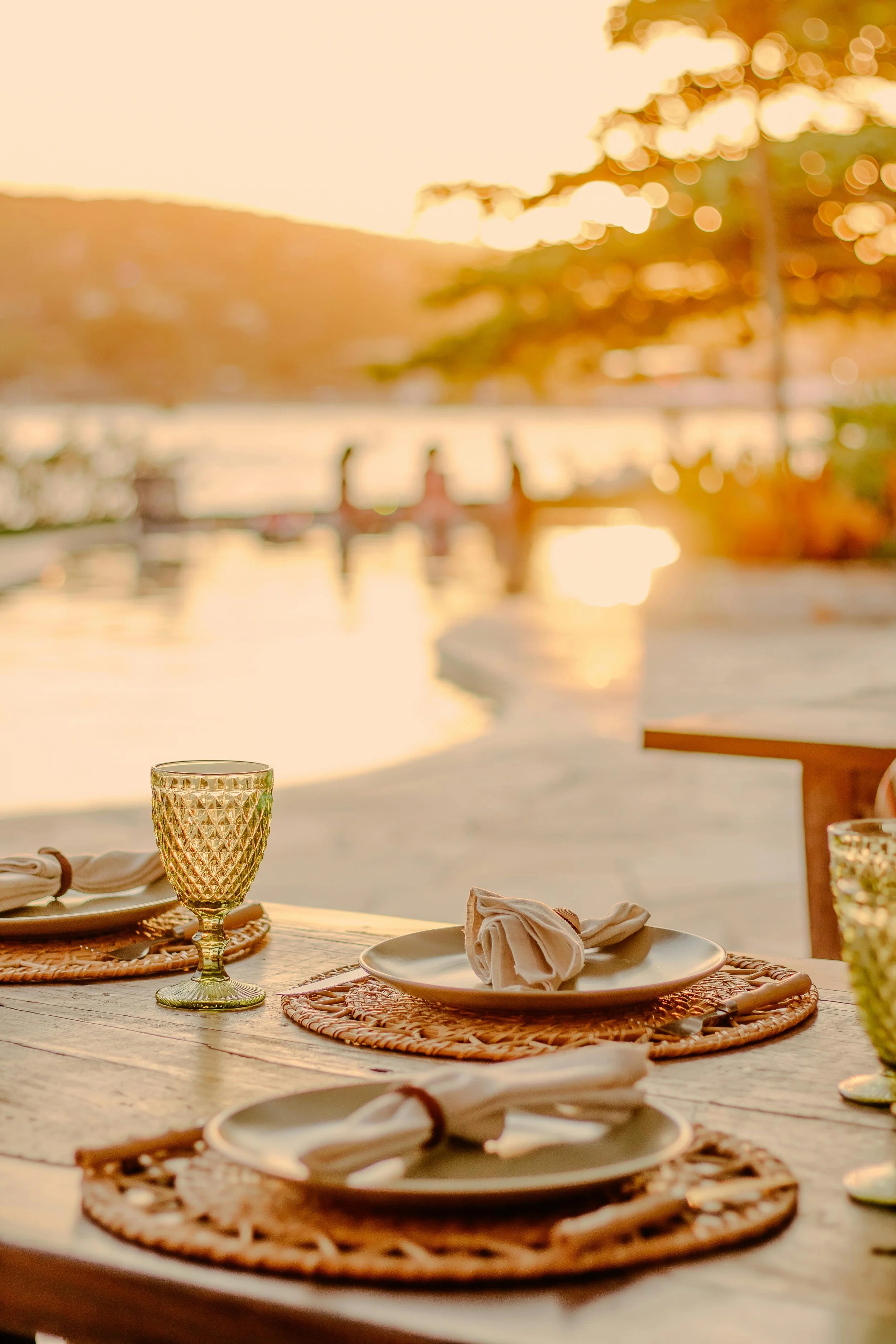 A table set for an outdoor meal with plates, napkins, and glasses, overlooking a river and trees in a sunset scene.