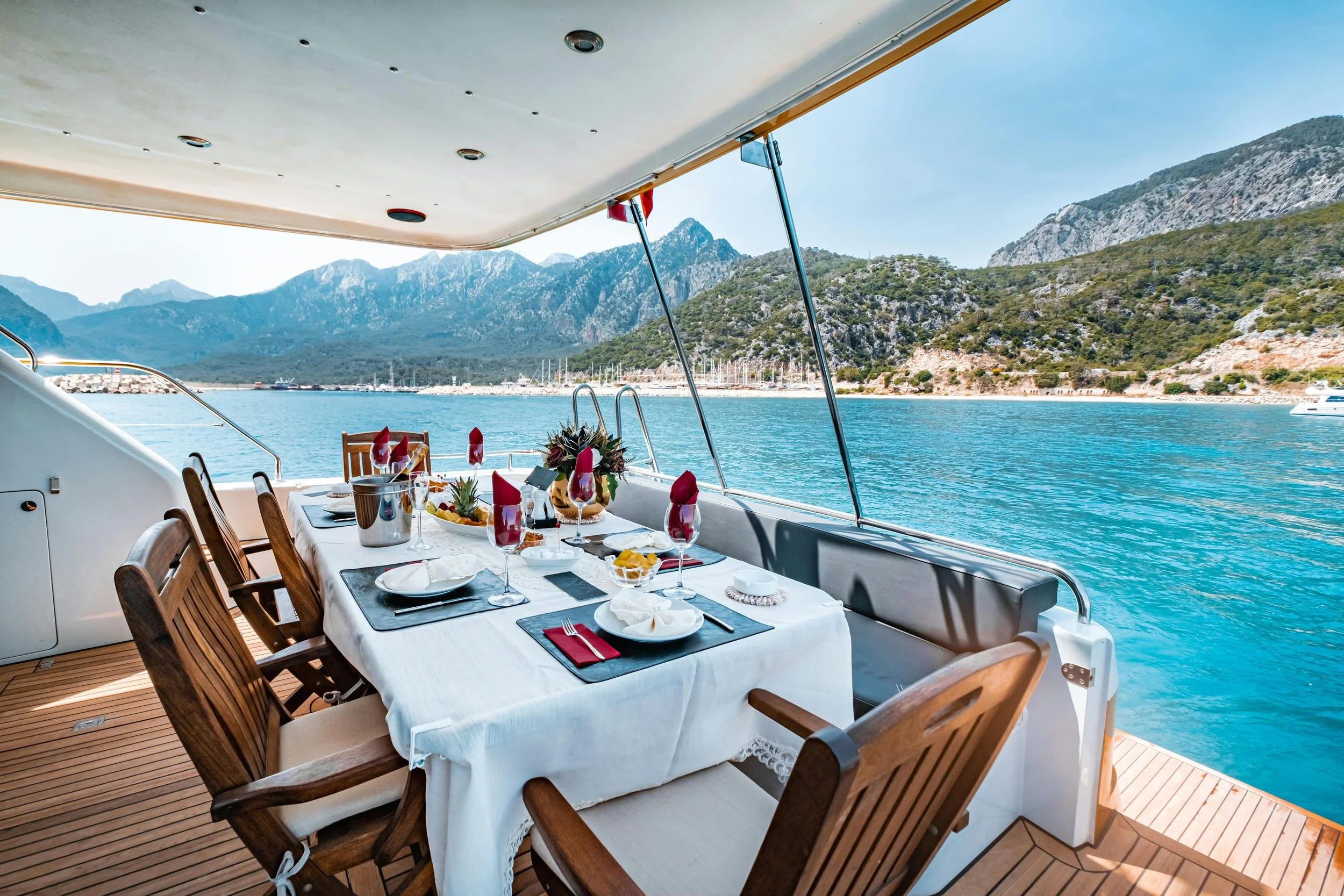 A dining table set for a meal on a yacht, overlooking a scenic mountain landscape with water and sailboats in the background.
