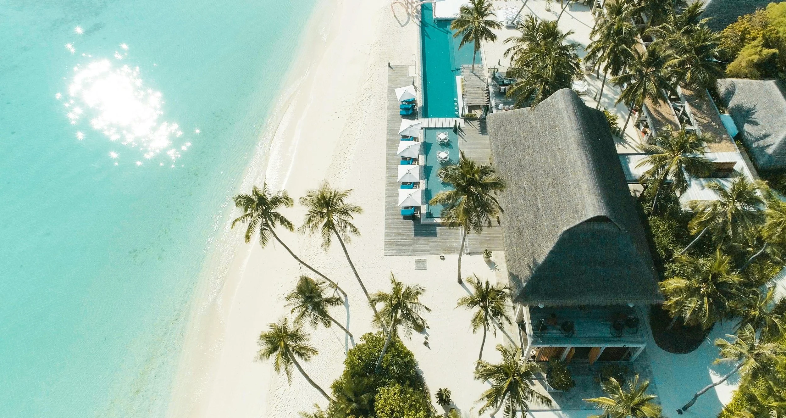 Aerial view of a beach resort with turquoise water, white sandy beach, palm trees, a pool, and thatched-roof buildings