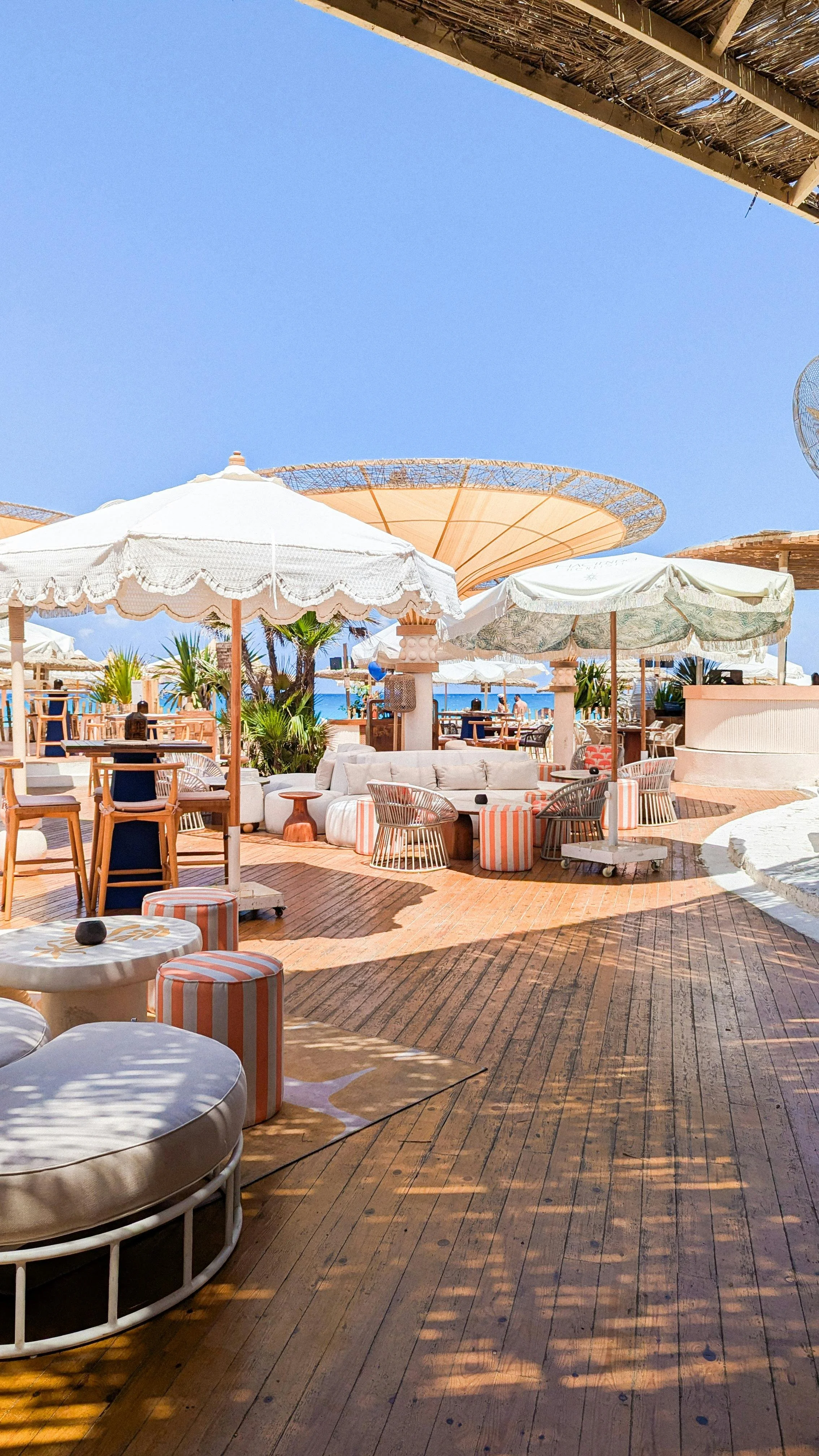 Beachside outdoor lounge area with umbrellas, seating, and palm trees, overlooking the ocean under a clear blue sky.