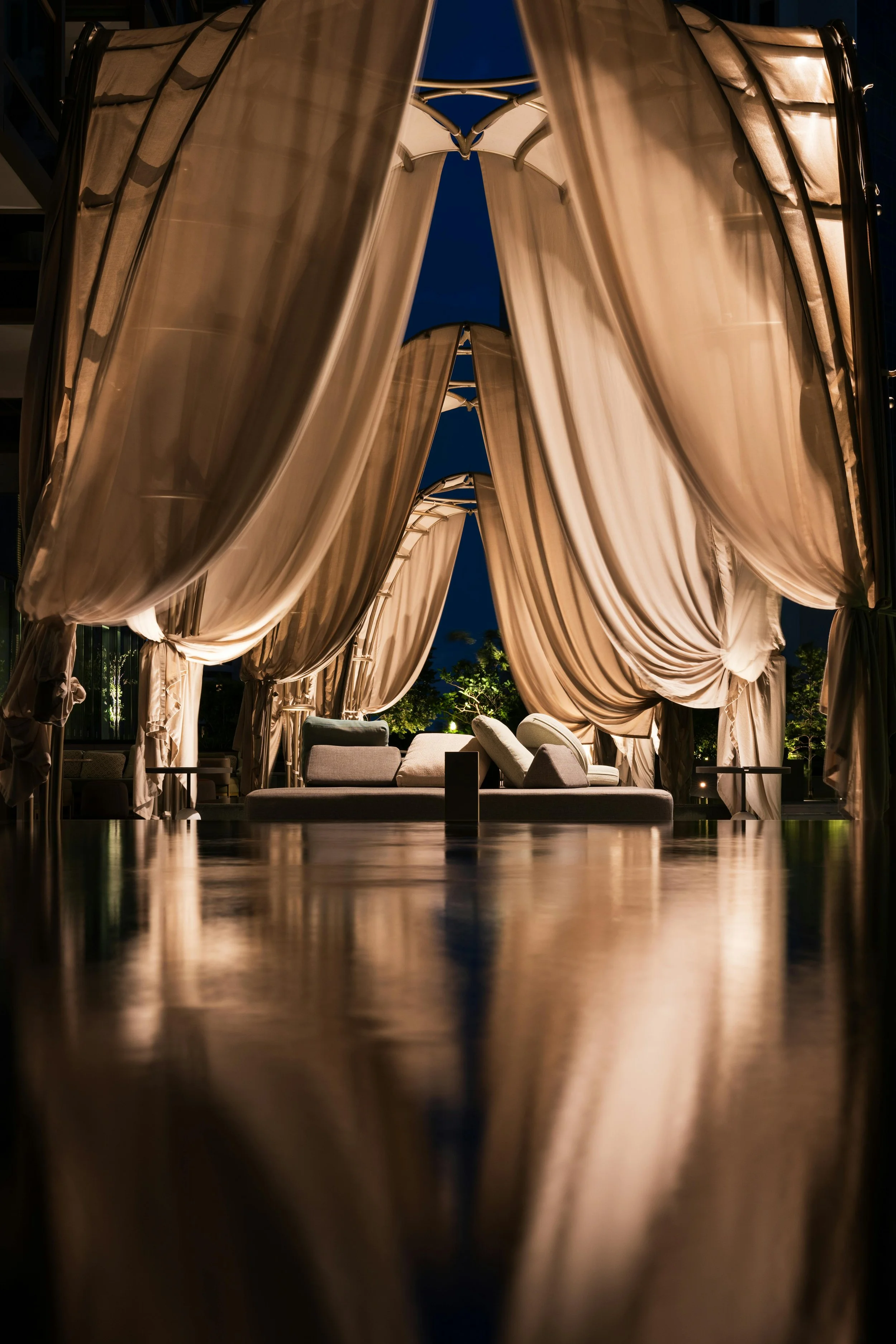 Nighttime outdoor lounge area with beige curtains and cushioned seating, illuminated softly with warm lighting, surrounded by greenery and trees.