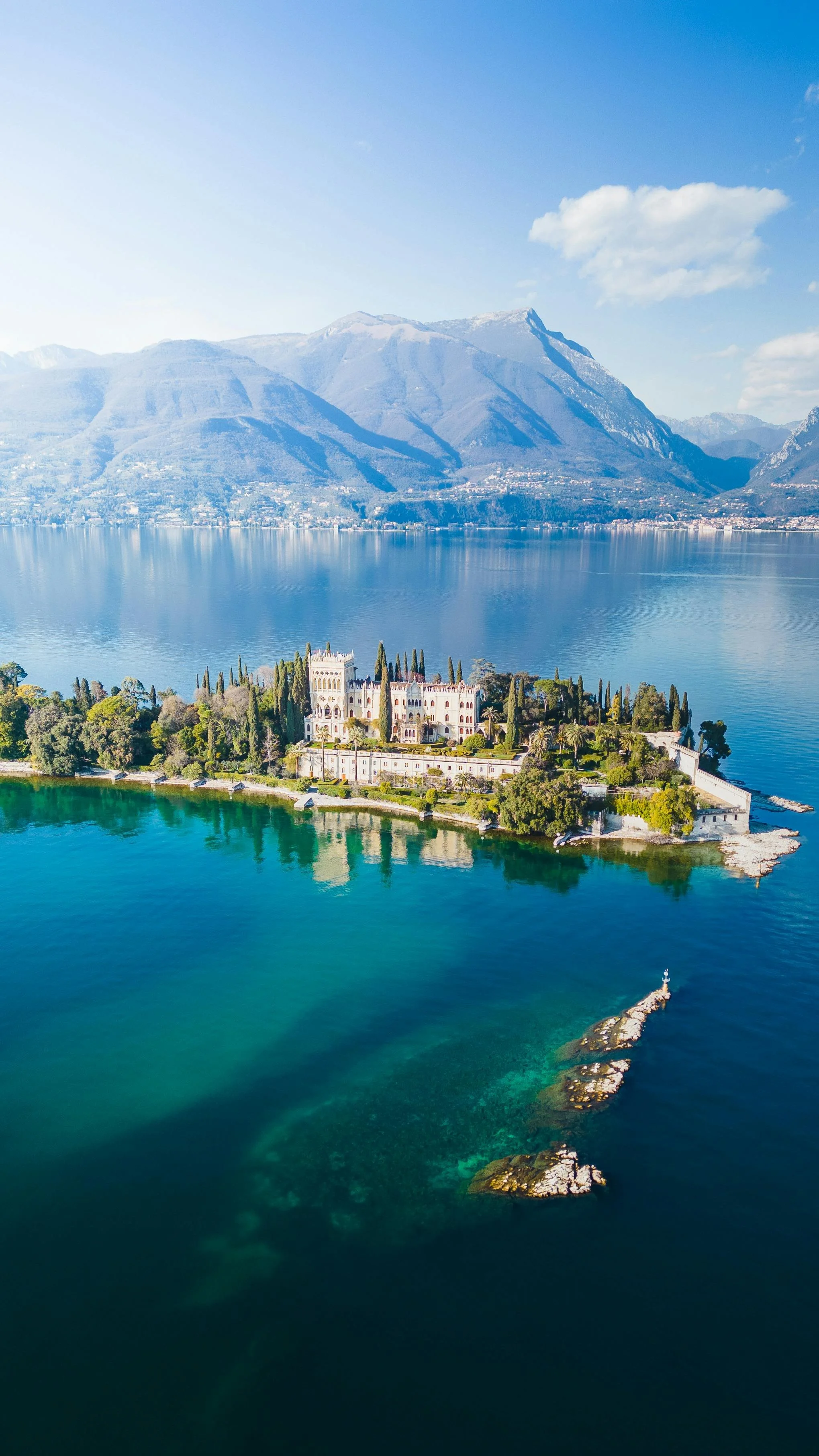 Aerial view of a castle on an island in a lake with snowy mountains in the background and a blue sky.
