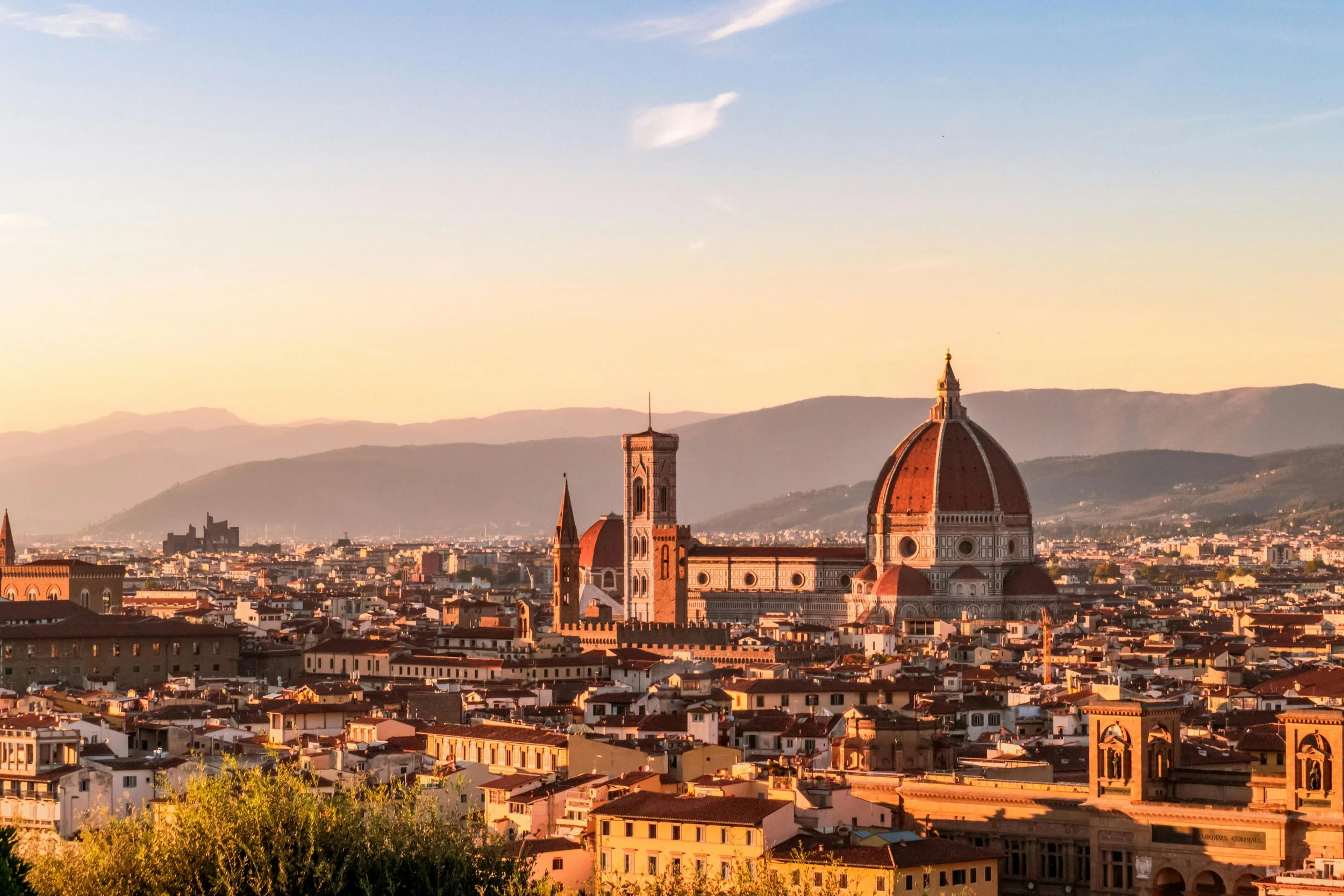 Sunset over Florence, Italy, with the Florence Cathedral's dome and bell tower visible among the city rooftops against a backdrop of mountains and a sky with pastel clouds.