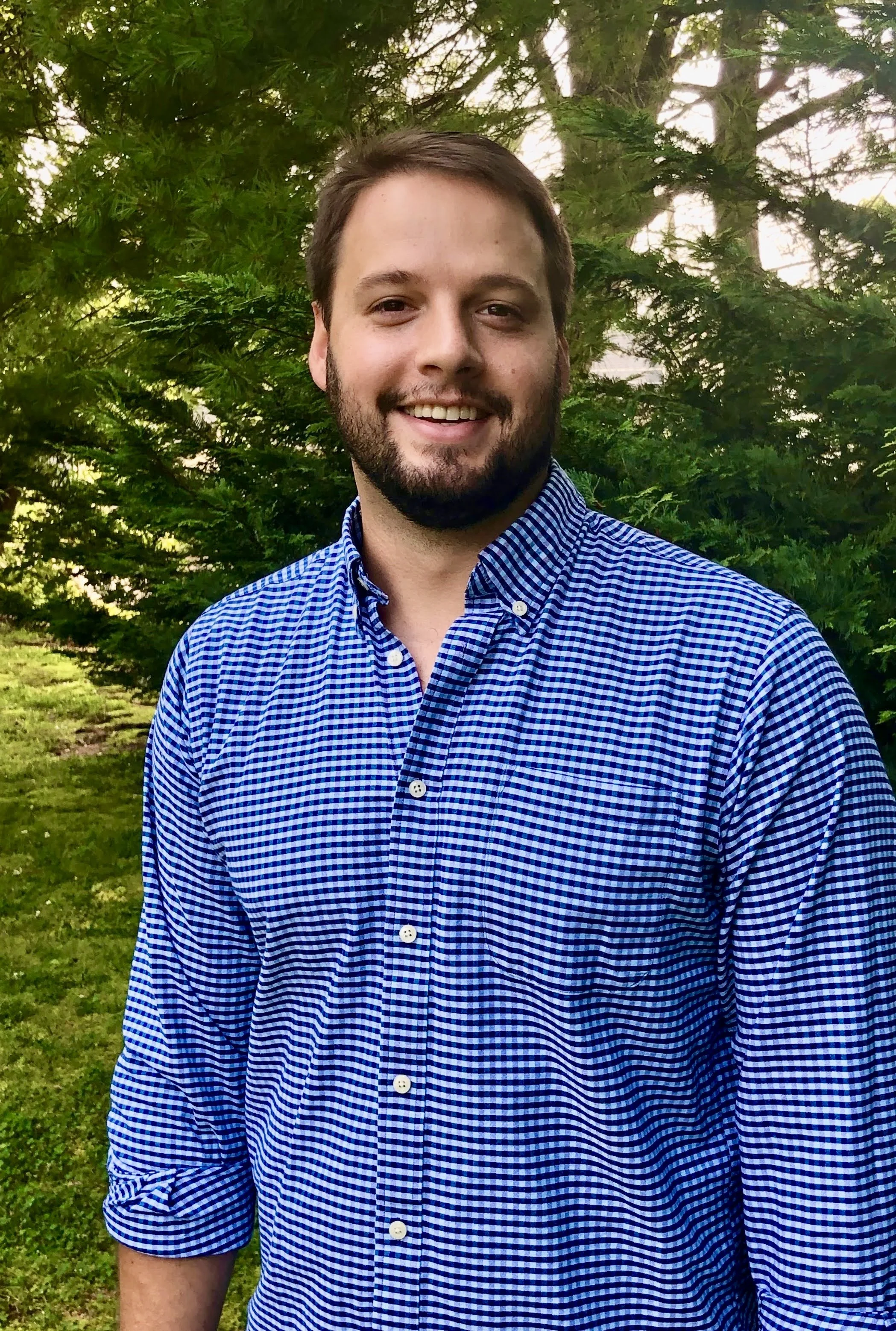 A man with a beard and short dark hair smiling outdoors in front of green trees, wearing a blue and white checkered button-up shirt.