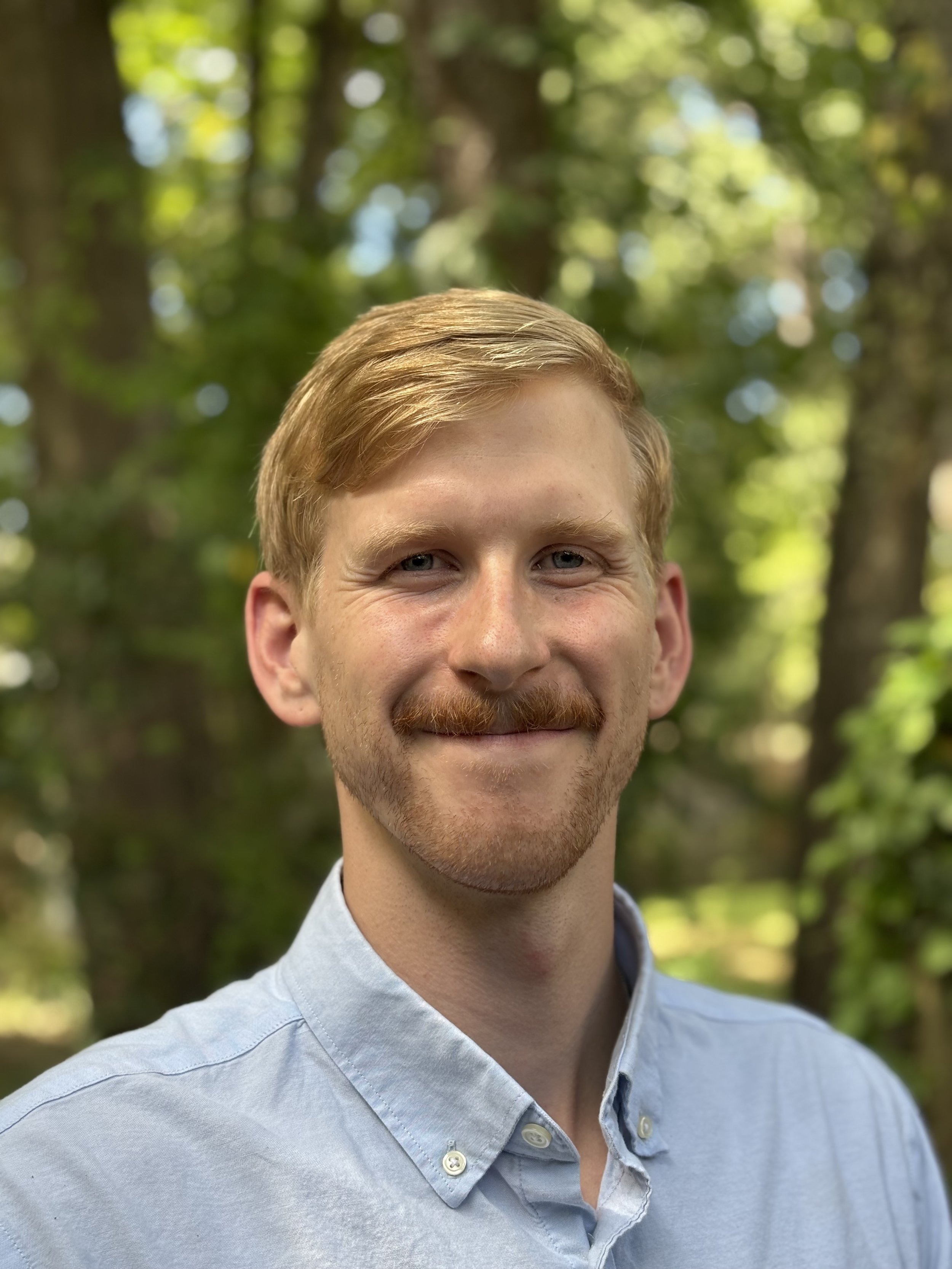 A young man with light red hair and a beard, wearing a light blue button-up shirt, smiling outdoors with a blurred green forest background.