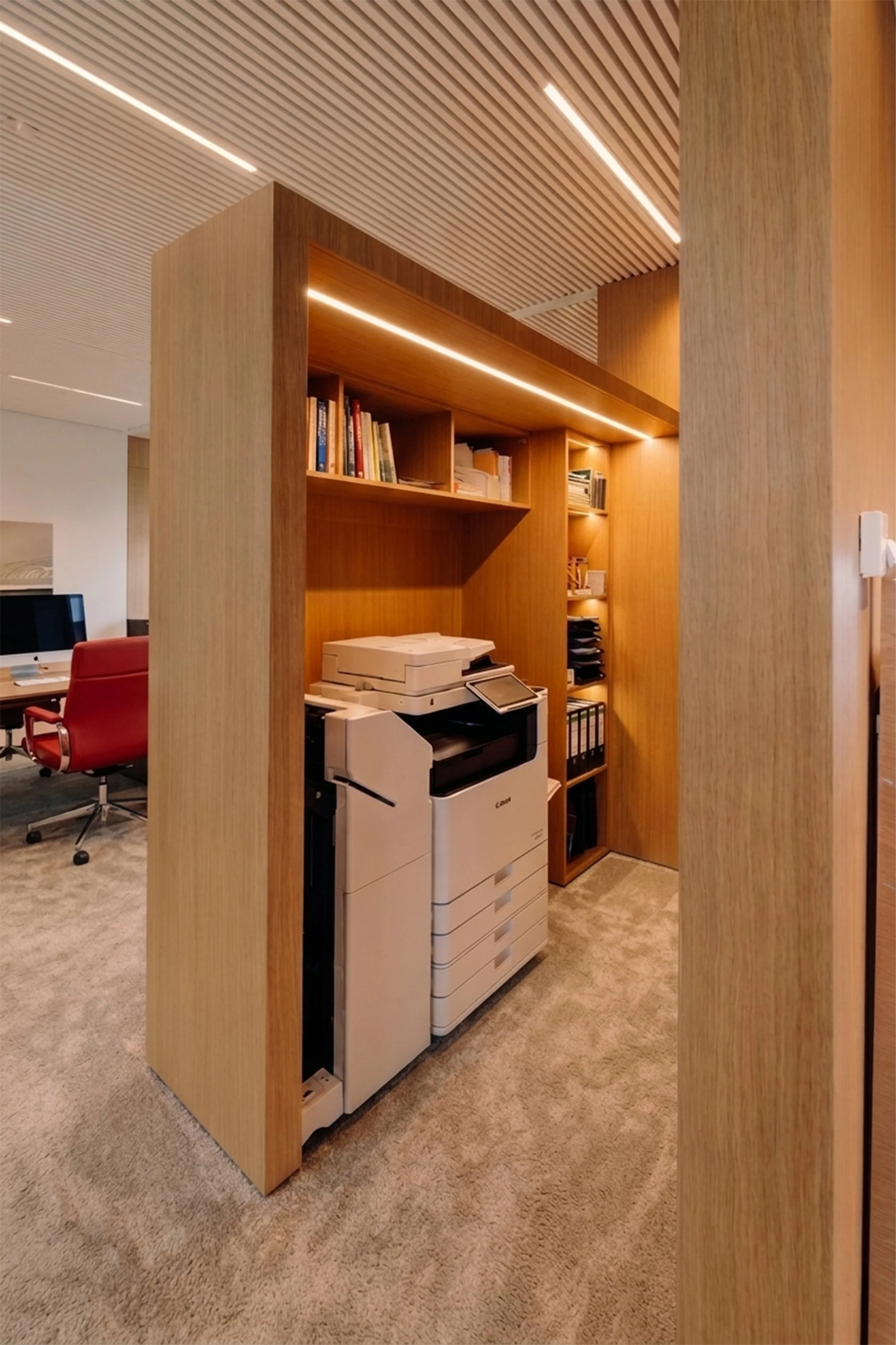 Un espace de bureau avec des meubles en bois, une imprimante multifonction, quelques livres sur une étagère en bois, un fauteuil rouge et une façade en bois visible. Le plafond est en lattes de bois avec des bandes lumineuses intégrées.