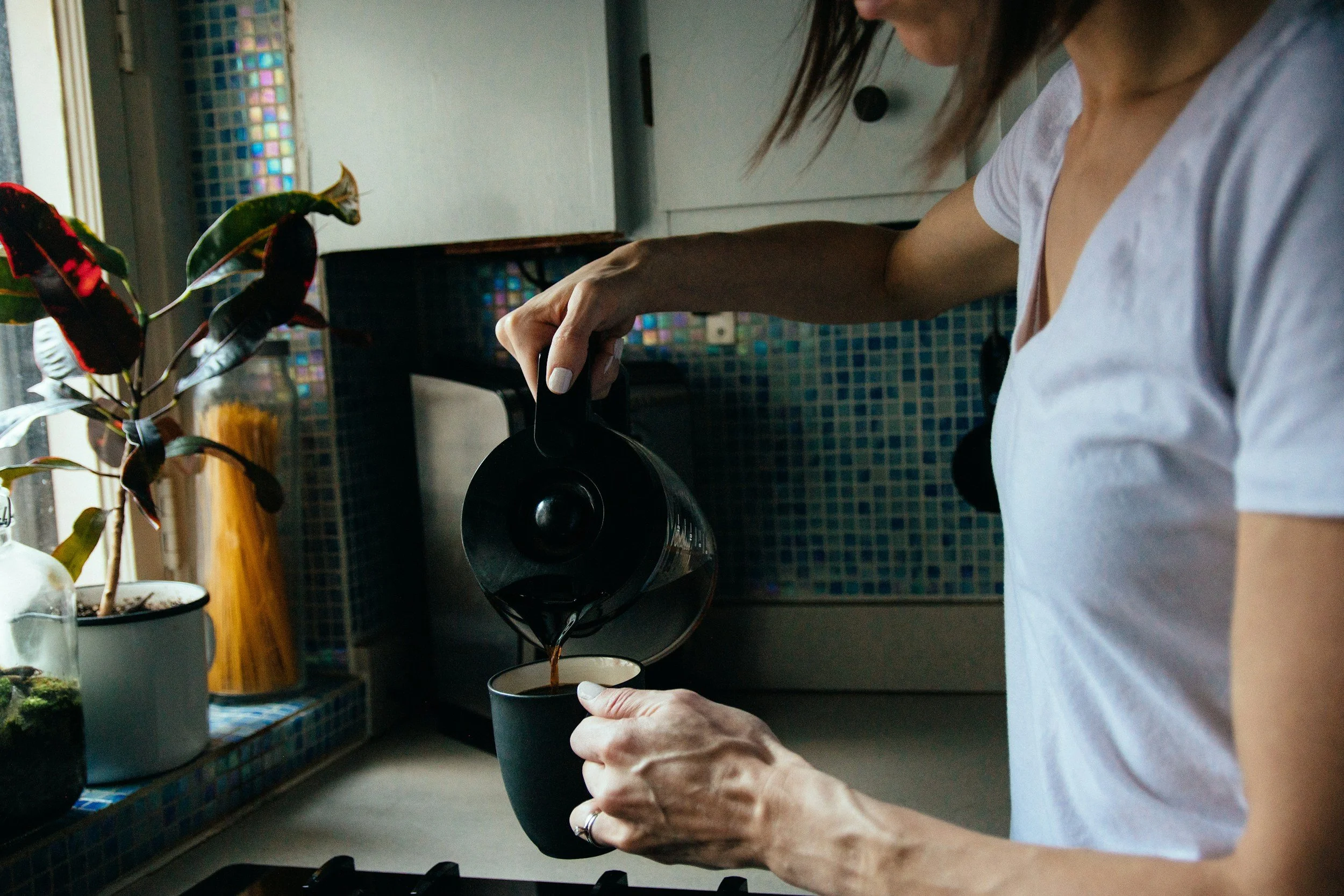 A woman pouring coffee into a mug in a kitchen with a potted plant and colorful mosaic tiles on the wall.