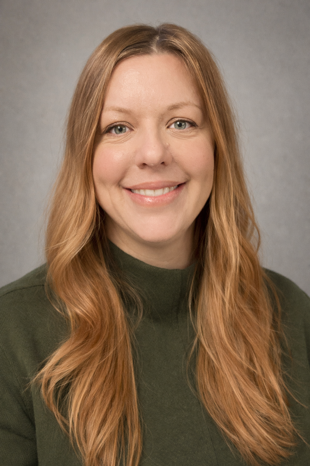 A woman with long red hair wearing a dark green top, smiling against a gray background.