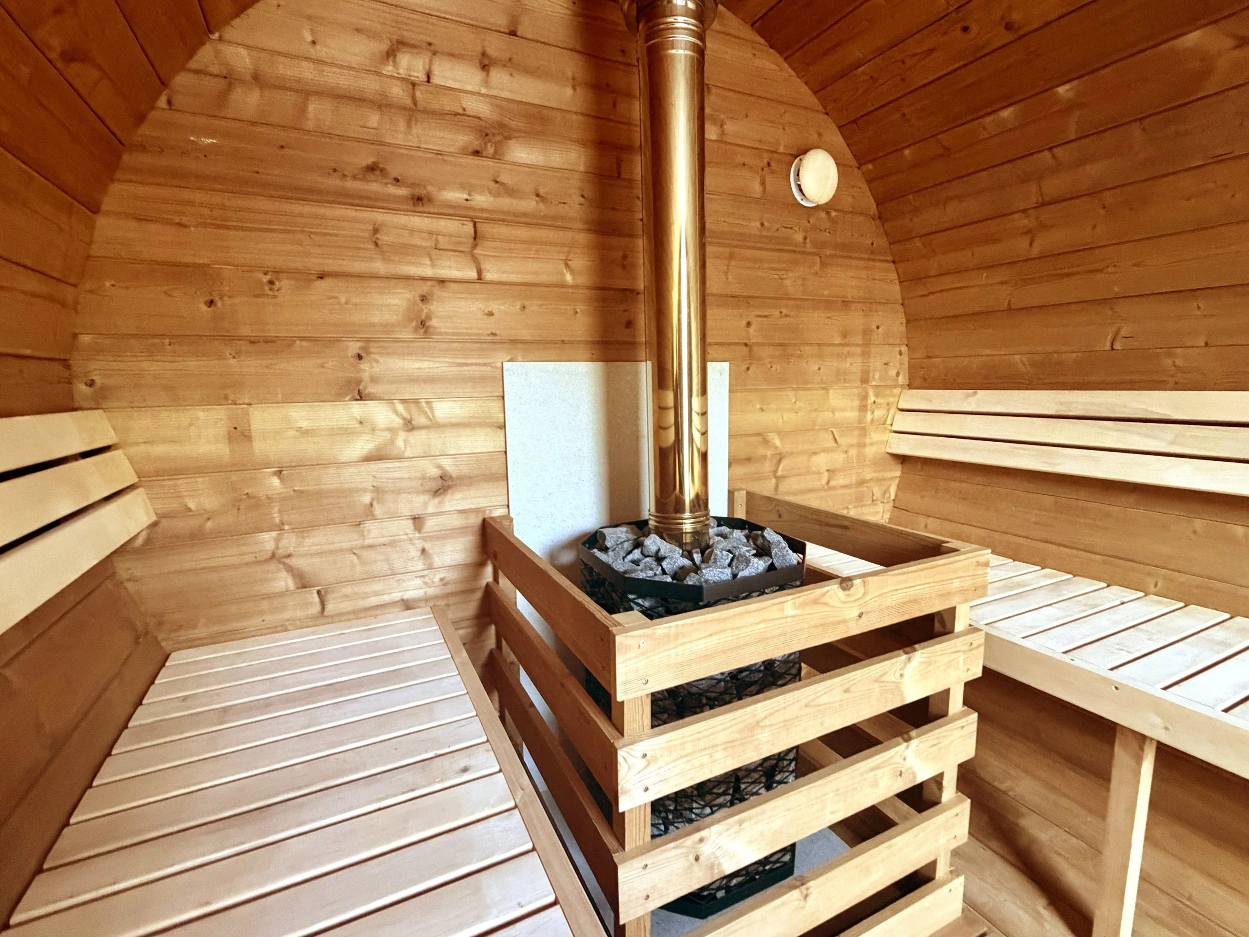 Interior of a wooden sauna with a stove in the center filled with rocks, surrounded by wooden benches on the sides and a circular vent on the wall.
