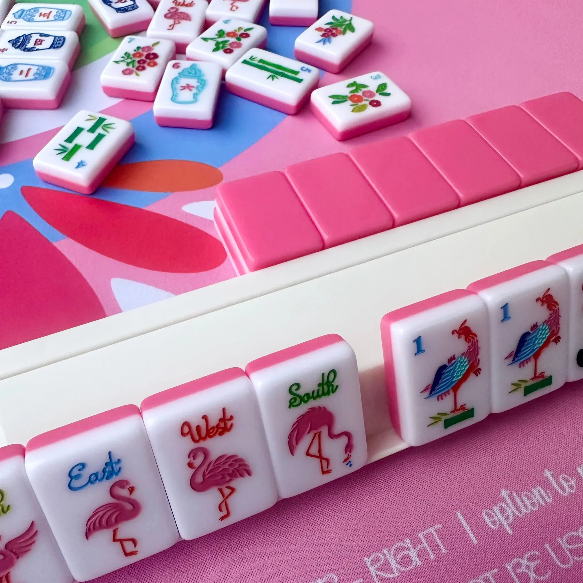 Close-up of a colorful Mahjong game set with tiles featuring floral, geographic, and animal designs on a pink, blue, and green background.