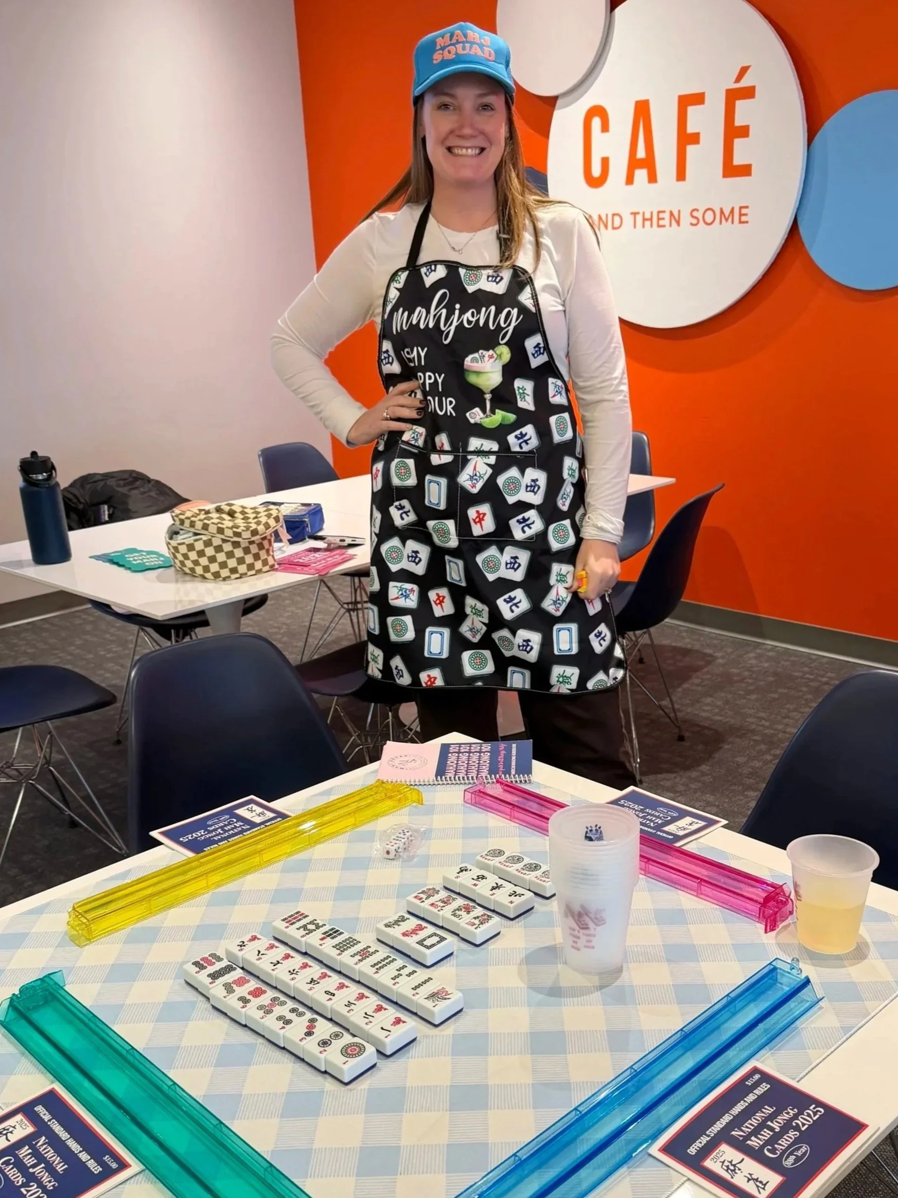 A woman standing in front of a table with mahjong tiles, wearing a mahjong-themed apron and a blue cap that says 'Mahjong Squad,' in a room with a red and white wall with a sign that says 'Café'.