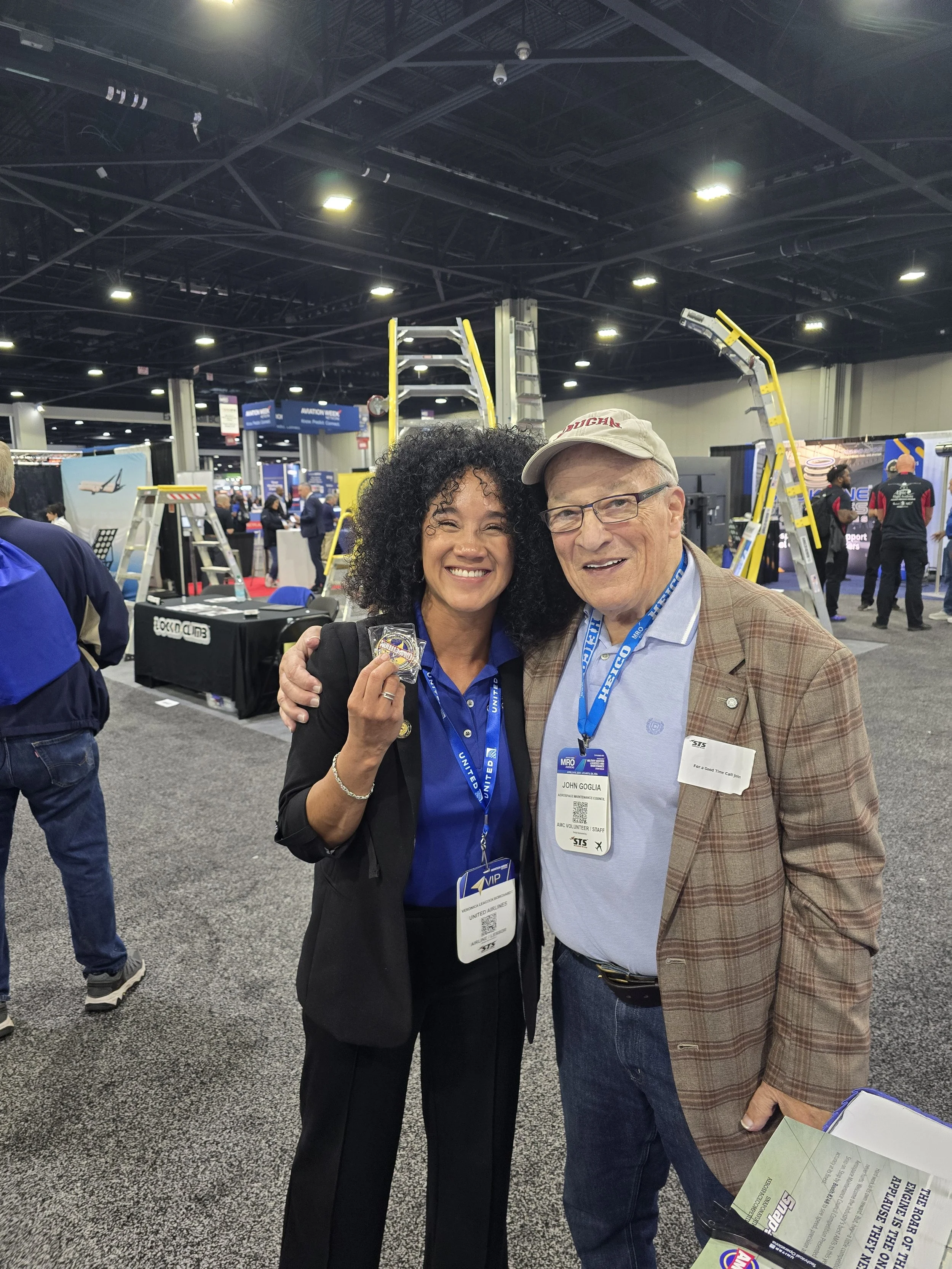 Two smiling individuals, a woman and a man, standing closely together, both wearing conference badges, in a busy exhibition hall with booths and ladders visible in the background. women in aircraft maintenance. Aircraft maintenance leadership
