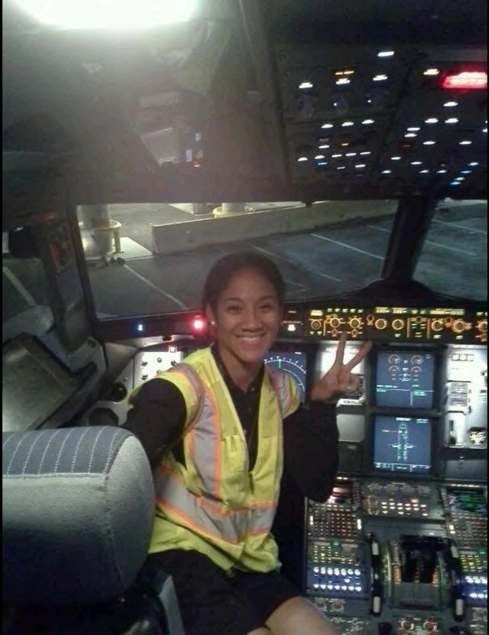 A woman smiling and making a peace sign inside the cockpit of an airplane, with numerous controls and screens visible around her.