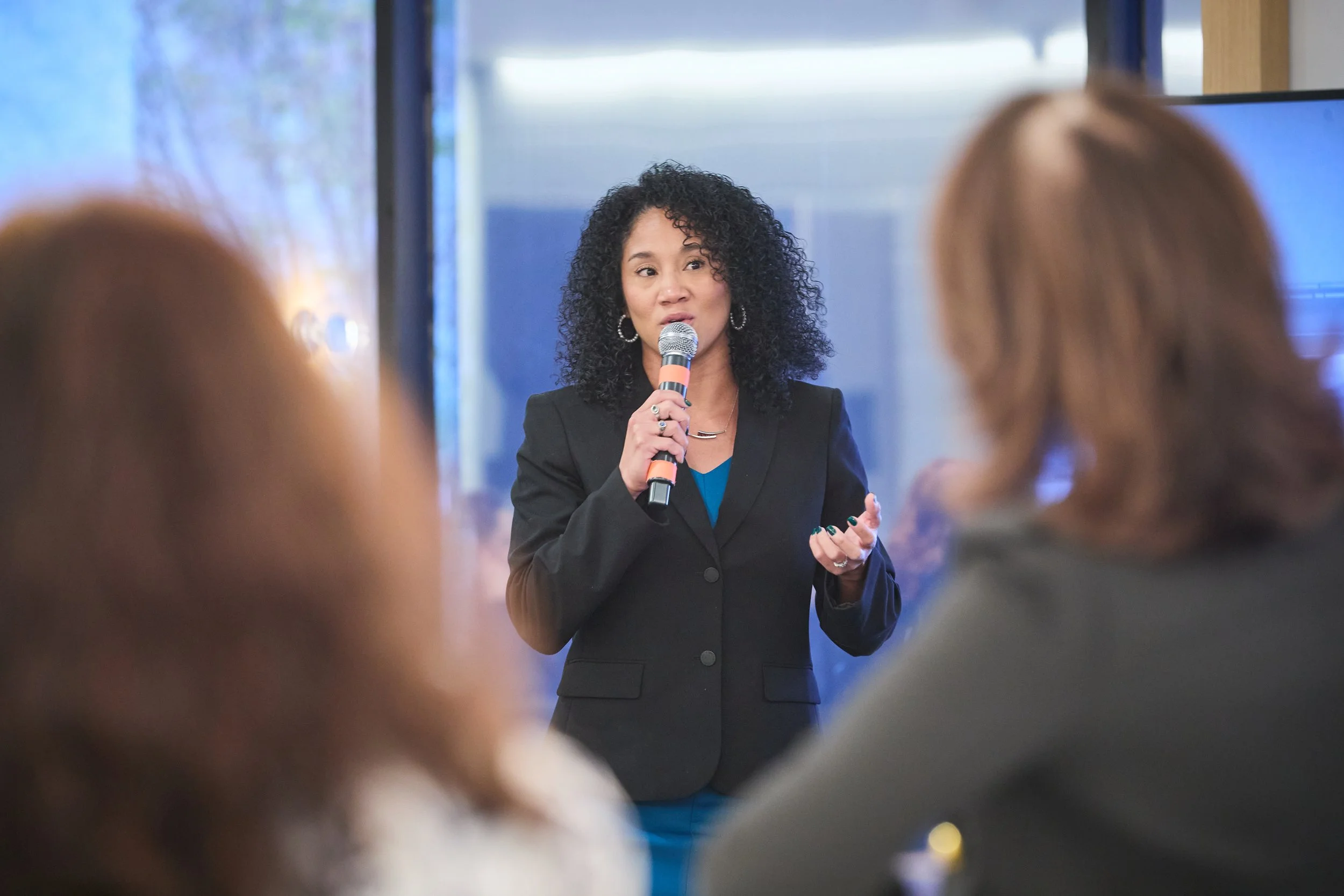 A woman with curly black hair wearing a black blazer and blue top speaking into a microphone during a presentation or speech. Women in aicraft maintenance
