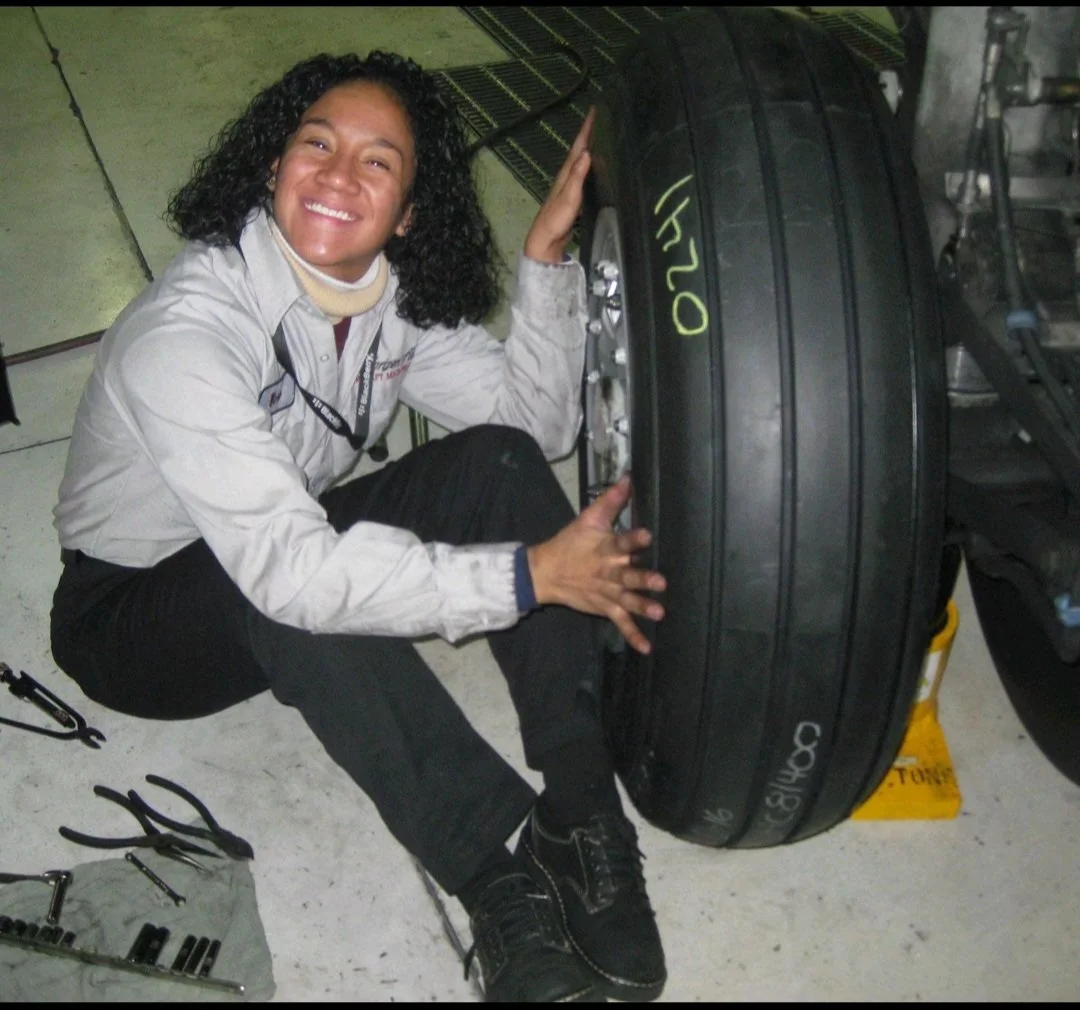A woman with curly black hair in work uniform kneeling next to a large black tire, smiling at the camera in a garage or workshop.