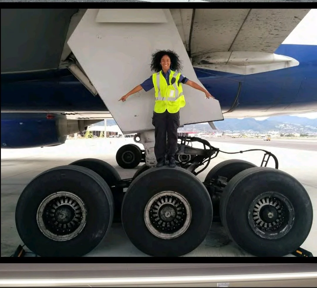 A woman in a yellow safety vest standing on the wheels of a large airplane at an airport. Women in aircraft maintenance