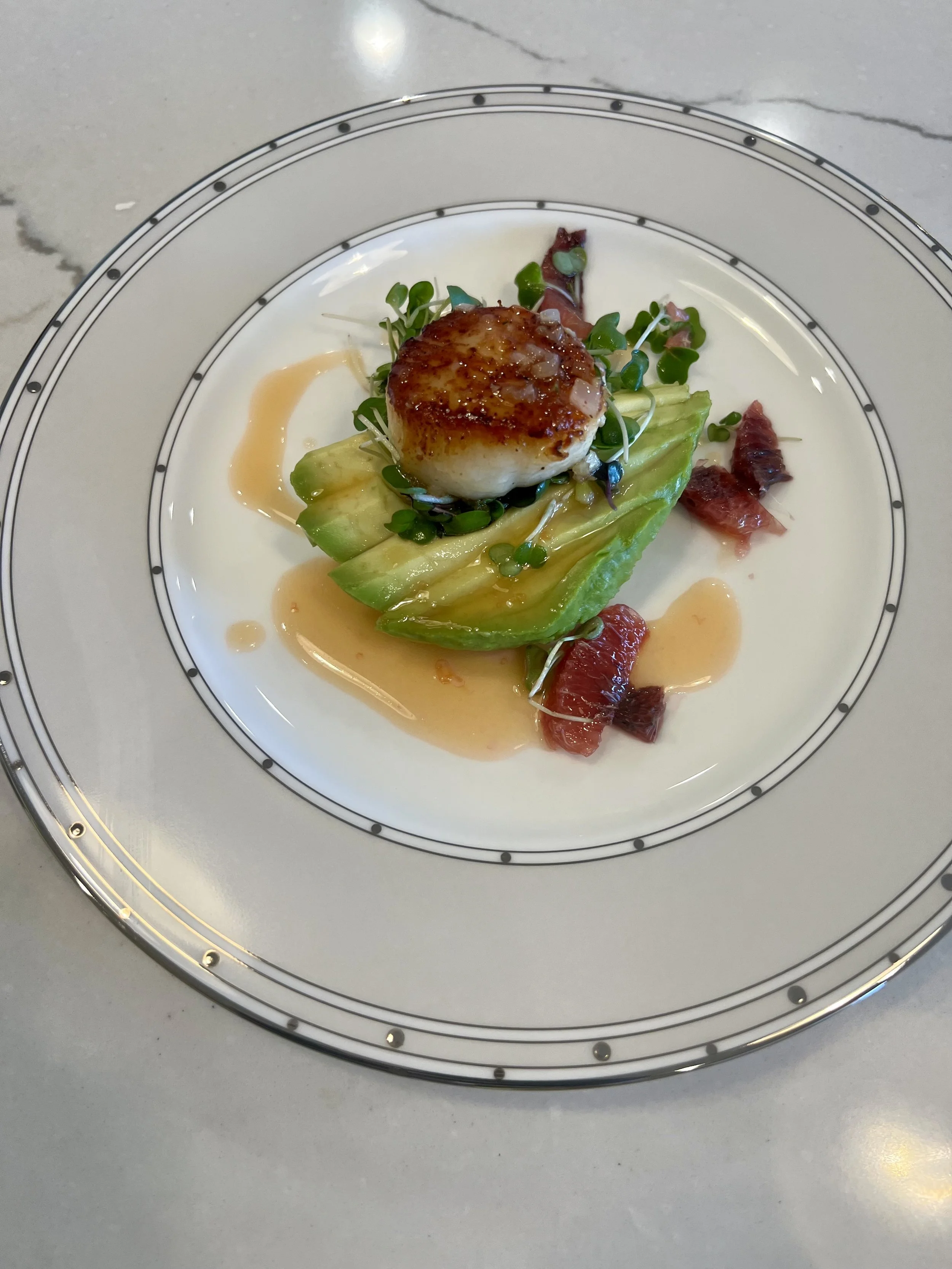 A plated gourmet dish featuring a seared scallop on a bed of avocado slices, microgreens, bacon pieces, and a light sauce.