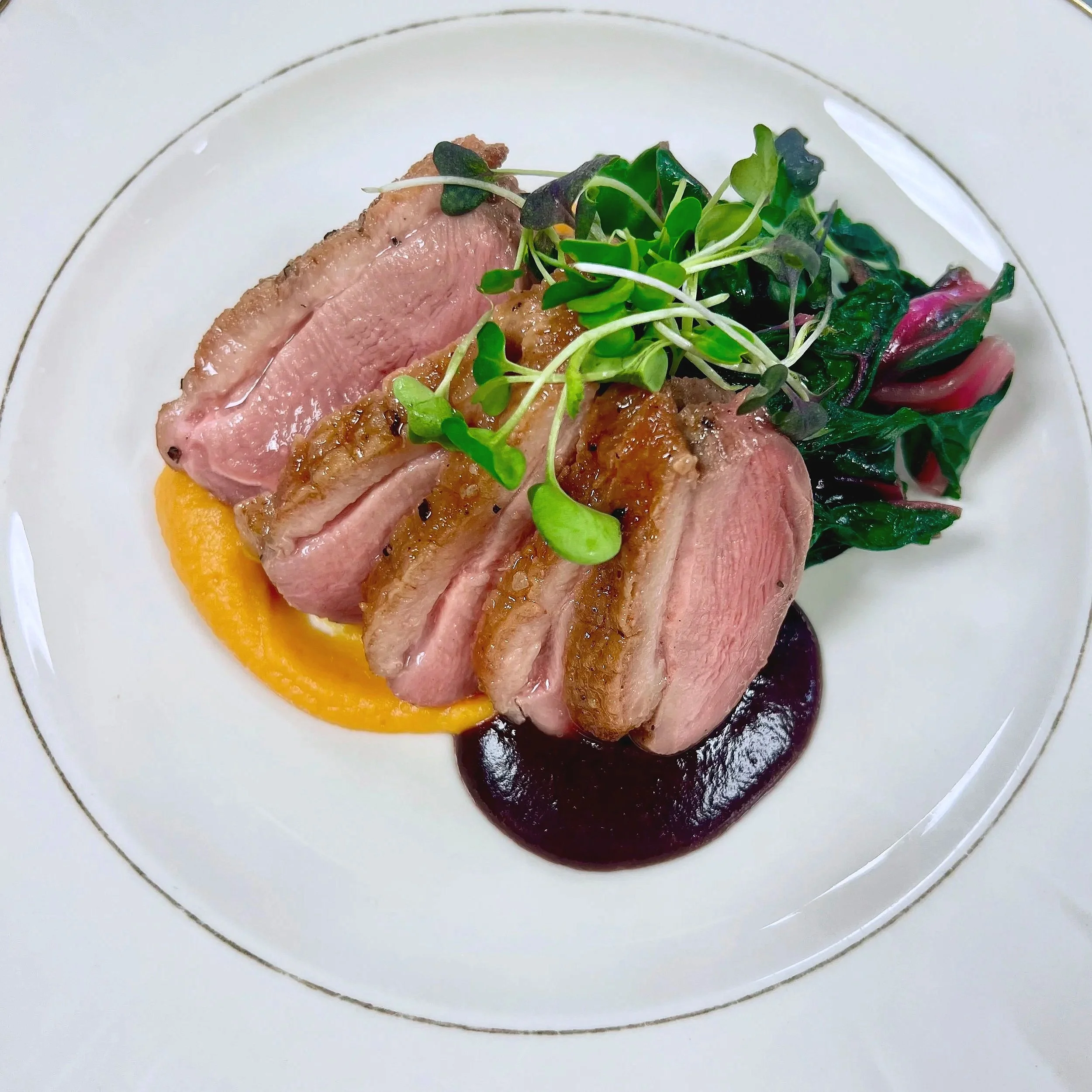 Seared duck breast sliced and plated with mango puree, berry sauce, and microgreens on a white plate.