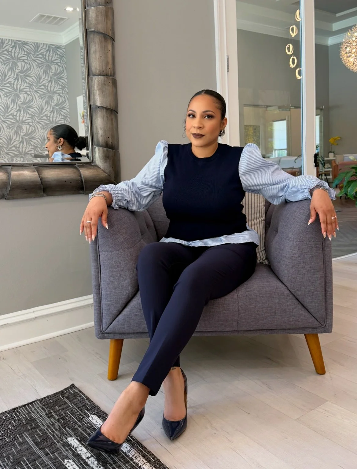 A woman sitting on a gray armchair with wooden legs inside a modern living room. She is wearing a navy blue and light blue striped blouse with puffed sleeves, black pants, and black high heels. Her hair is pulled back, and she is wearing makeup and jewelry. In the background, there's a large mirror reflecting her profile, a decorative wallpaper, and a glass sliding door leading to another room with a chandelier and round hanging lights.