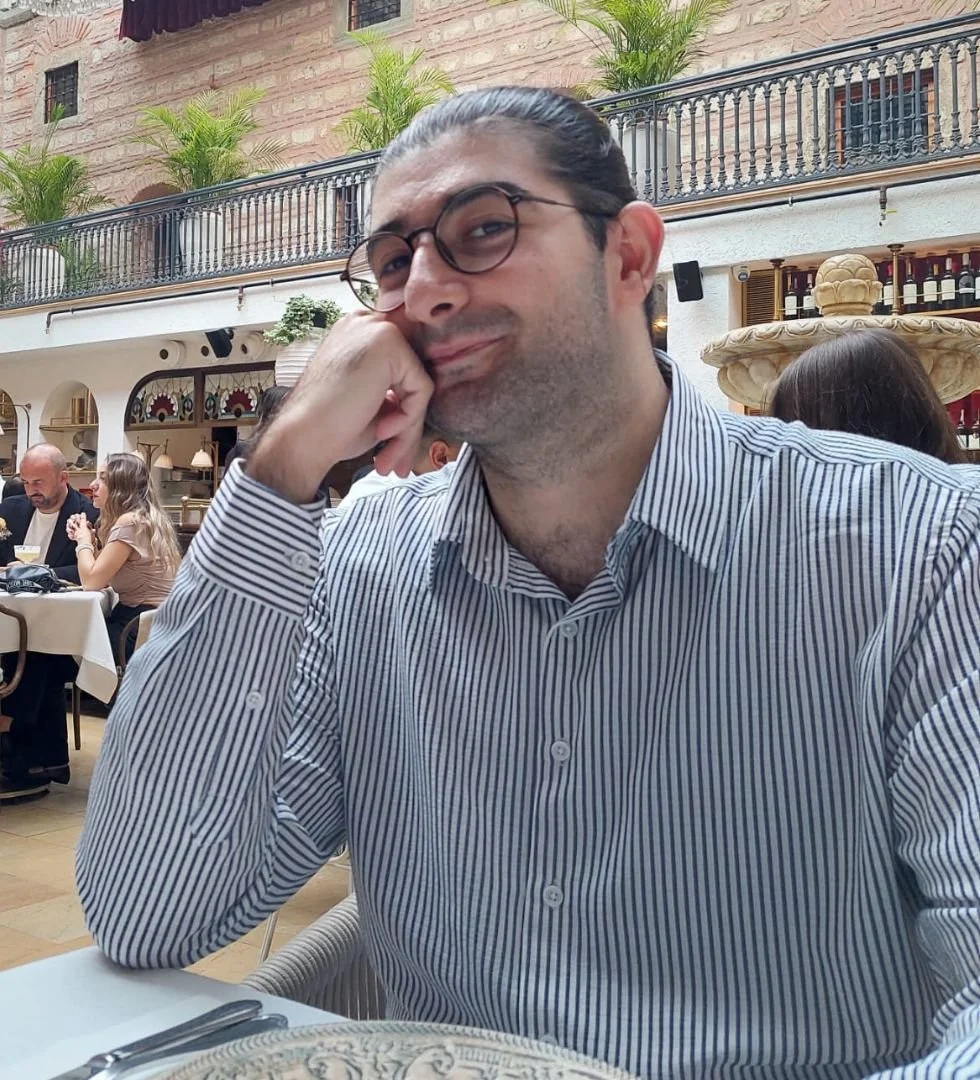 A man with glasses and a striped shirt sitting at a restaurant table, leaning his head on his hand, smiling at the camera. There are other diners and a decorative fountain in the background.