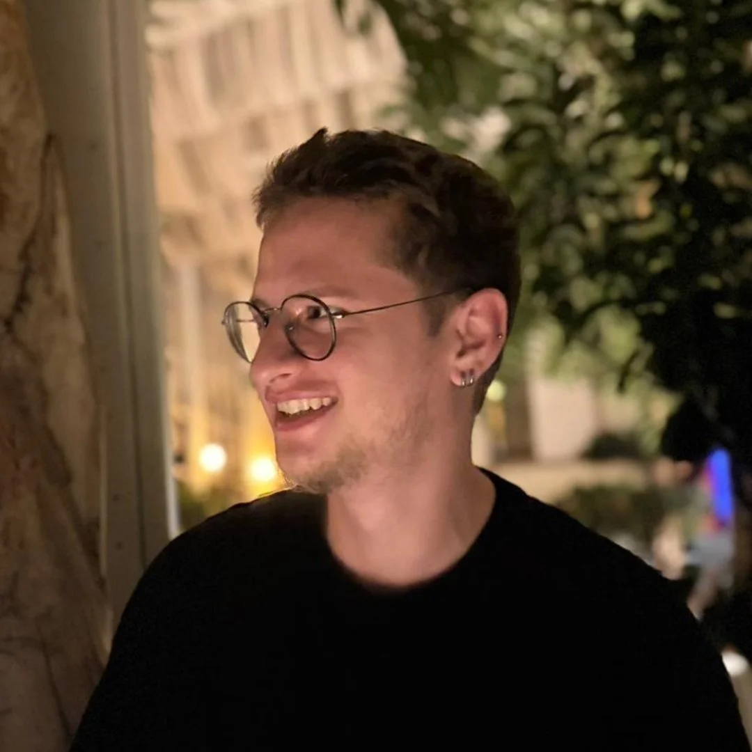 A smiling young man with glasses and earrings, standing outdoors in a nighttime setting with warm lighting and trees in the background.