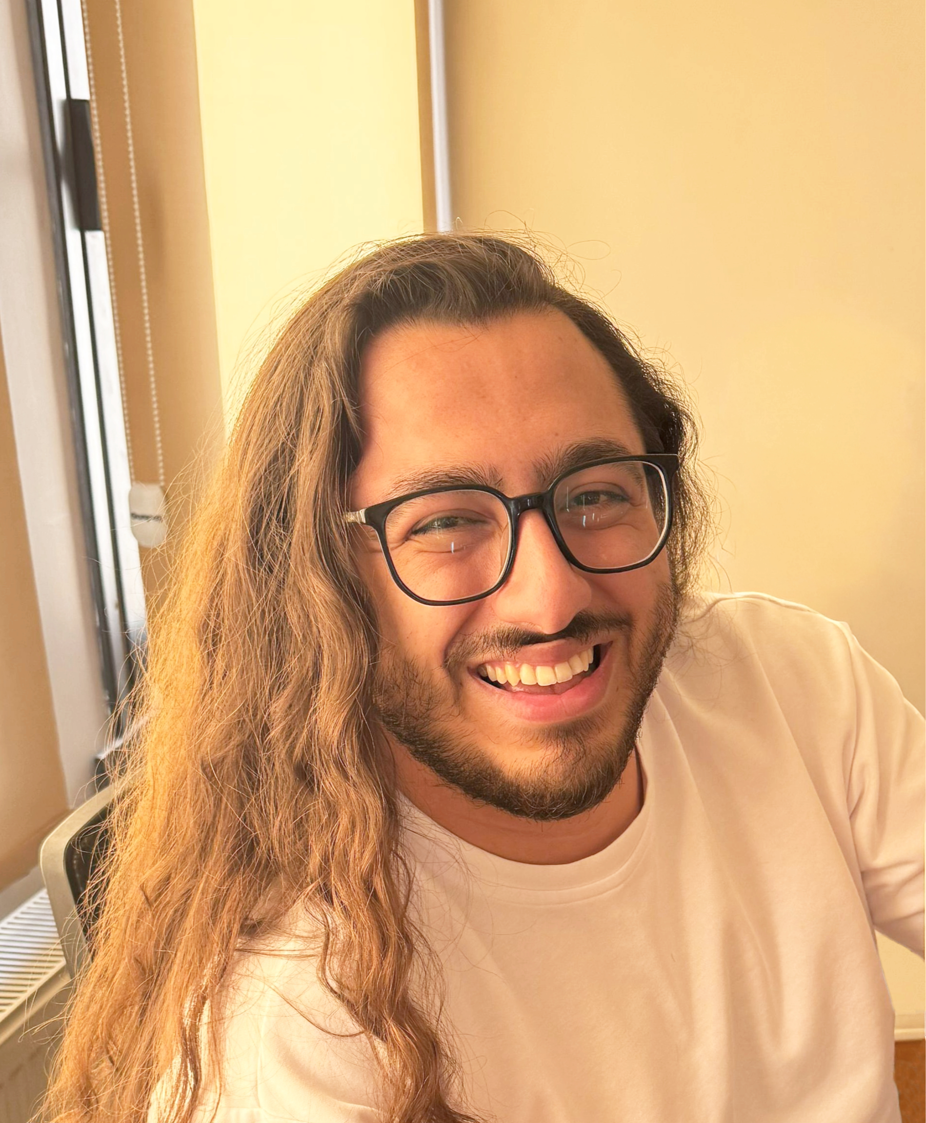 A man with long wavy hair wearing glasses smiling indoors near a window with beige walls.
