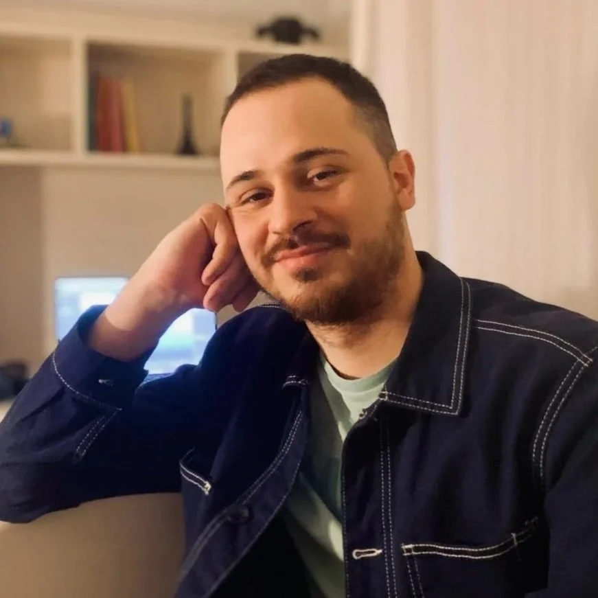 A man with short dark hair and a beard sitting indoors, resting his head on his hand, wearing a dark denim jacket and smiling.