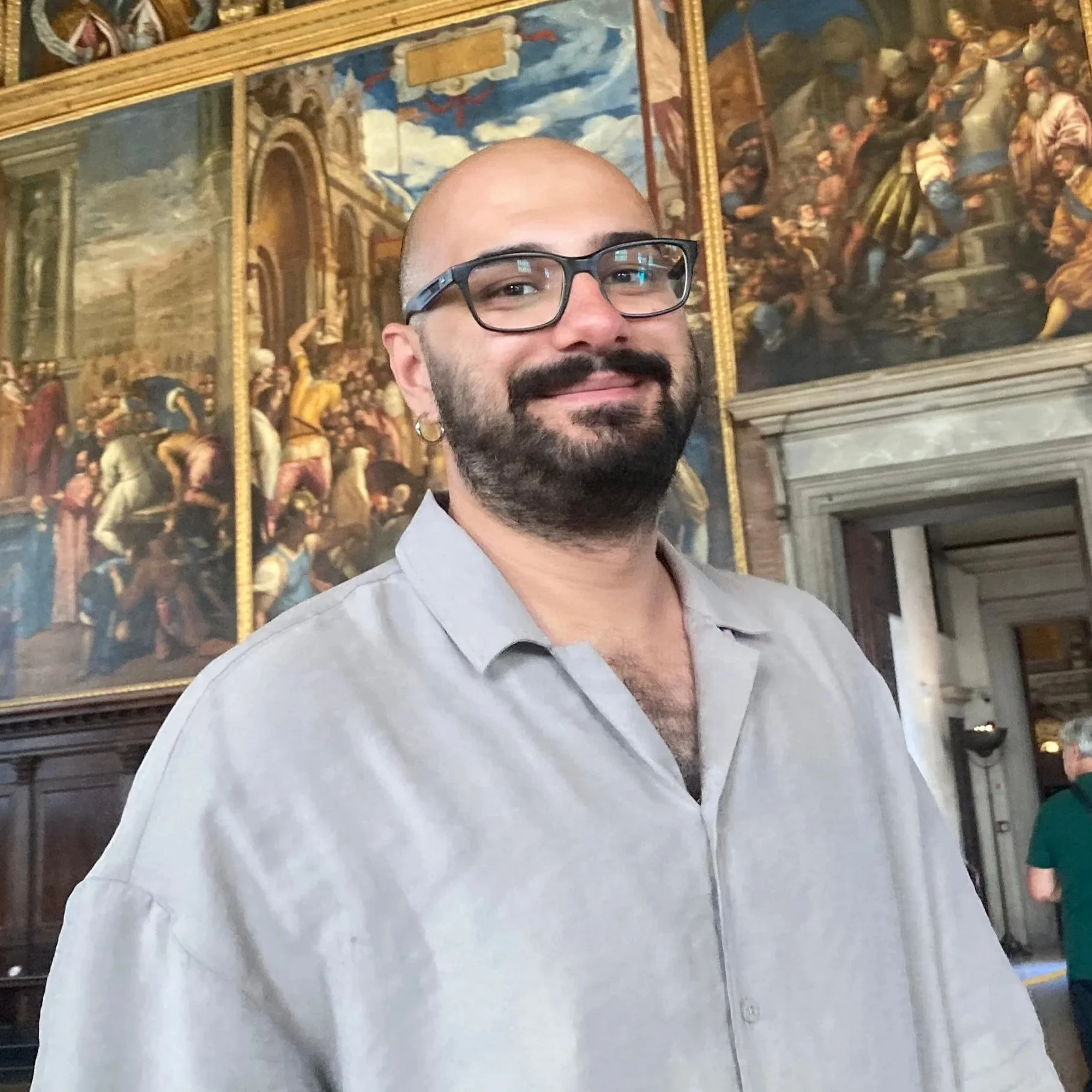 A man with a shaved head, glasses, and a goatee, wearing a light-colored shirt, standing in front of a large painting depicting a historical or religious scene inside a building with ornate decor.