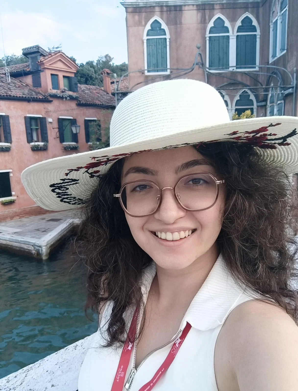 A young woman with glasses and curly hair smiling while standing near water, wearing a wide-brimmed hat and white sleeveless top, with colorful buildings in the background.