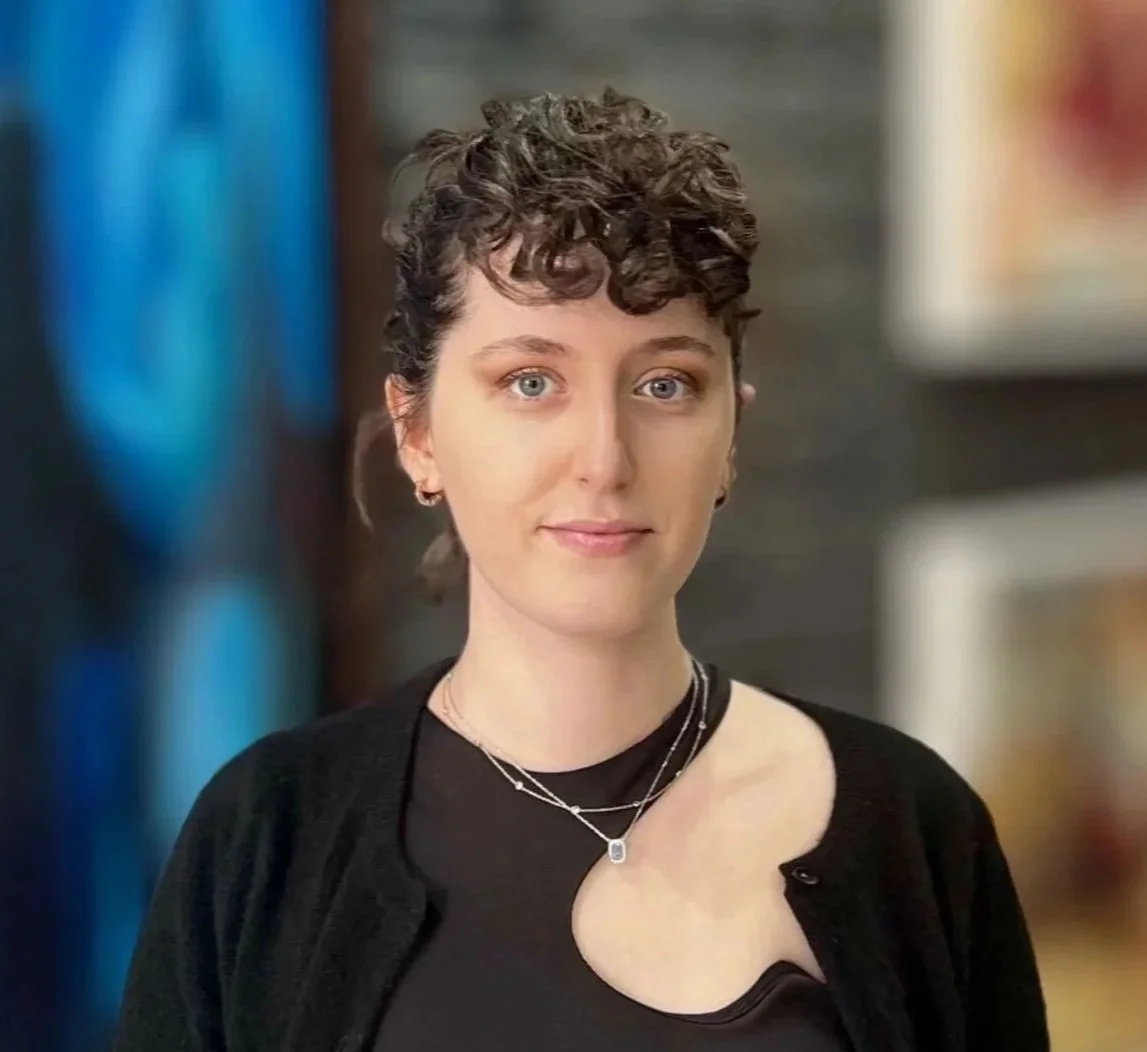 A woman with short, curly dark hair, light skin, and blue eyes, wearing a black top and a black cardigan, standing indoors with a blurred background.